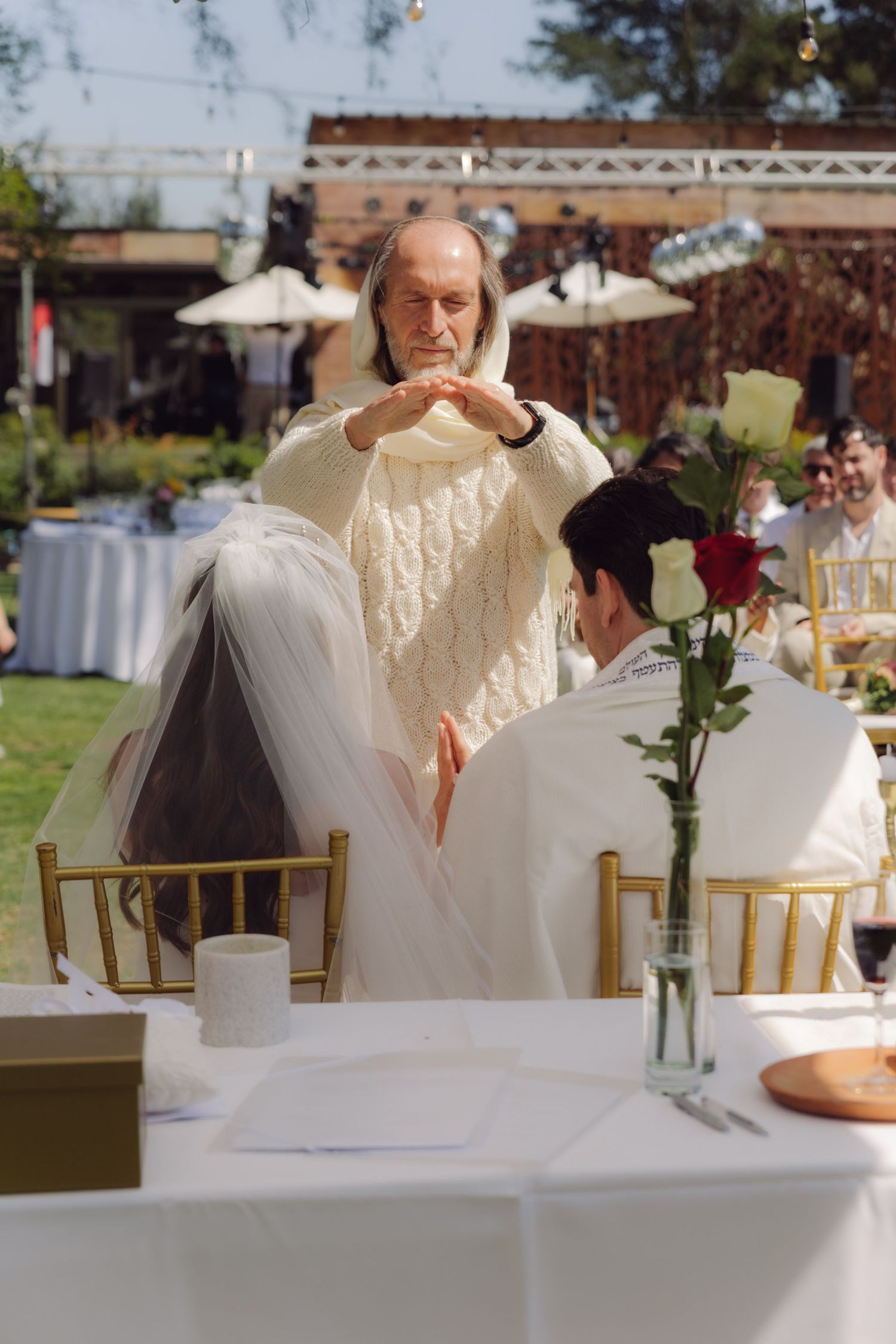 Elderly officiant in white robe performing an outdoor Jewish wedding ceremony for seated couple.