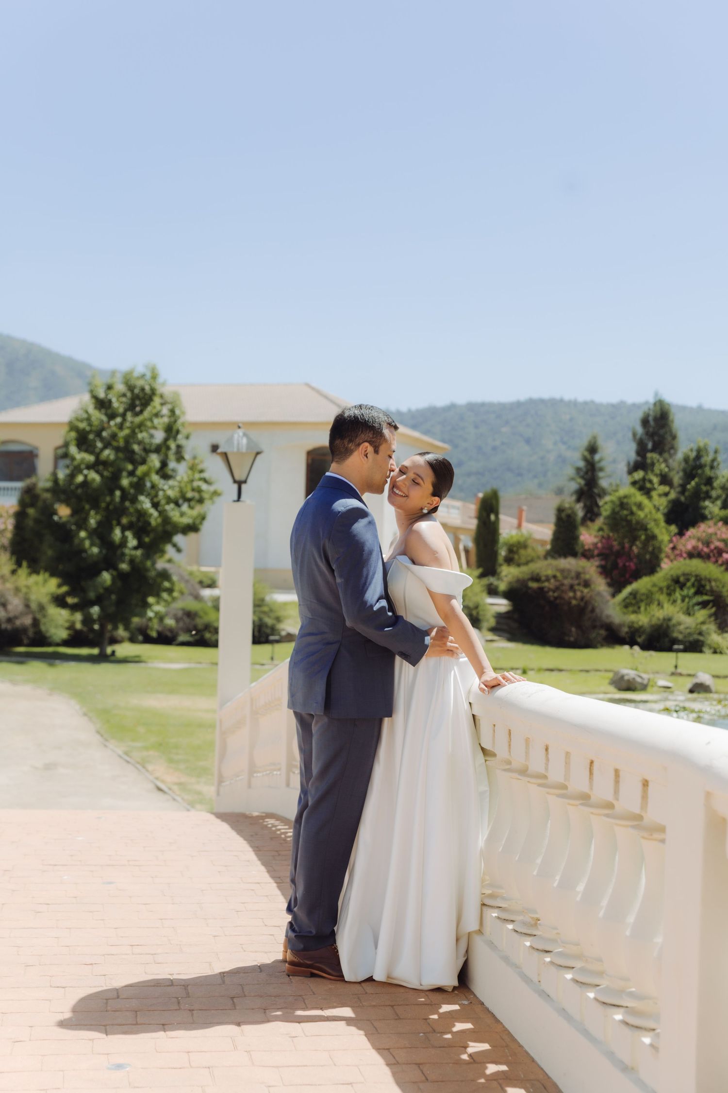 Bride and groom share a romantic embrace on a sunlit terrace with lush green gardens and mountains in the background.