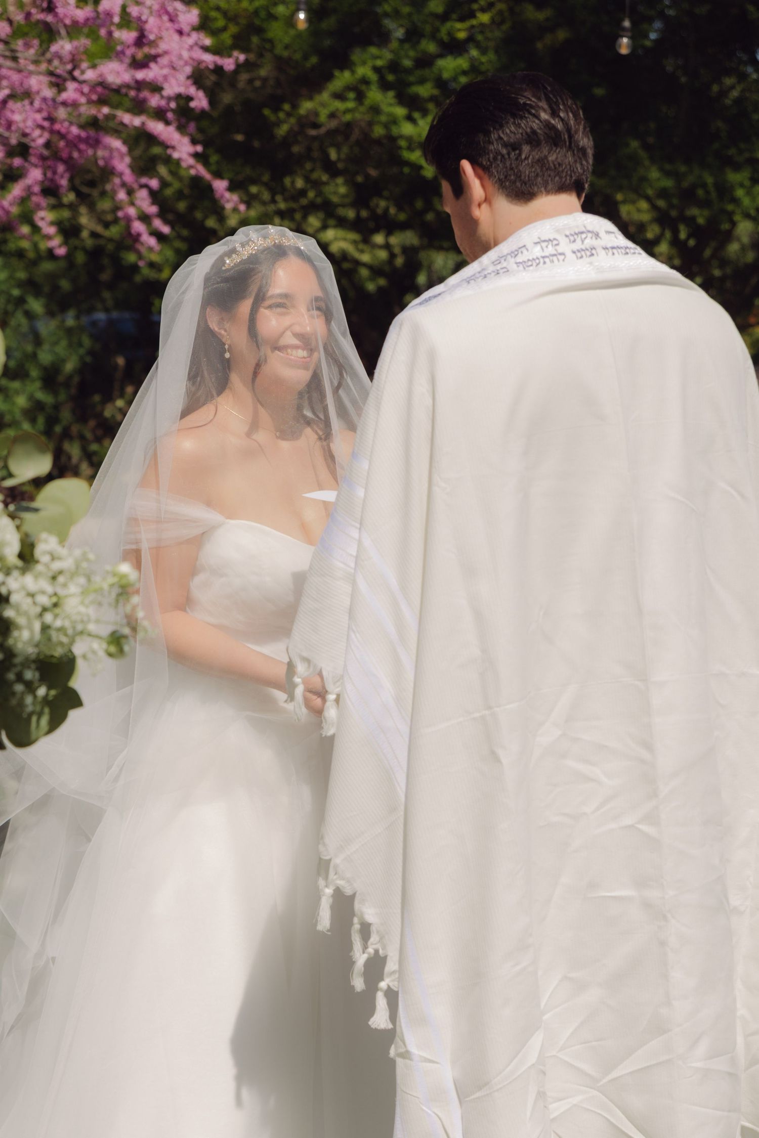 Smiling bride in white gown and veil holds hands with groom wearing a Jewish tallit outdoors.
