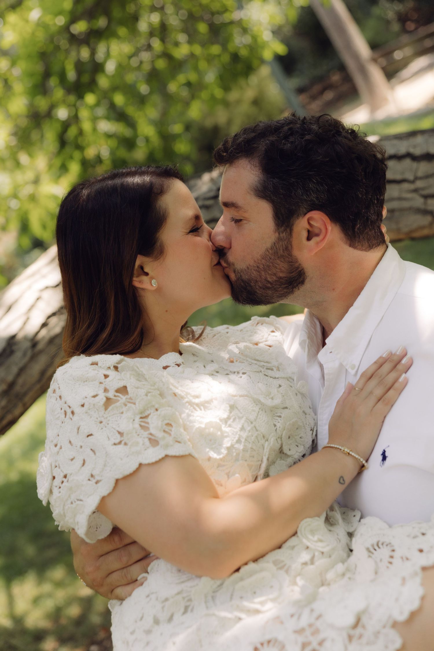 Couple shares a passionate kiss outdoors under green trees, woman wearing elegant white lace dress embracing man in white shirt.