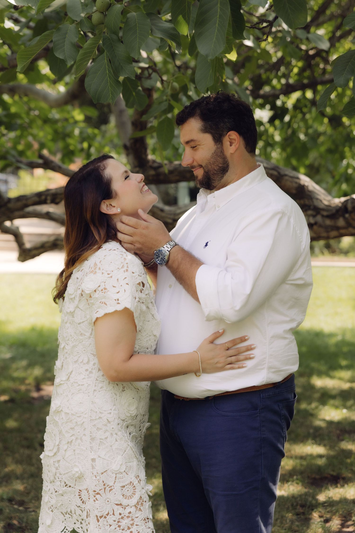 Couple shares a tender moment under a tree, man in white shirt gently holds womans face as she smiles up at him.