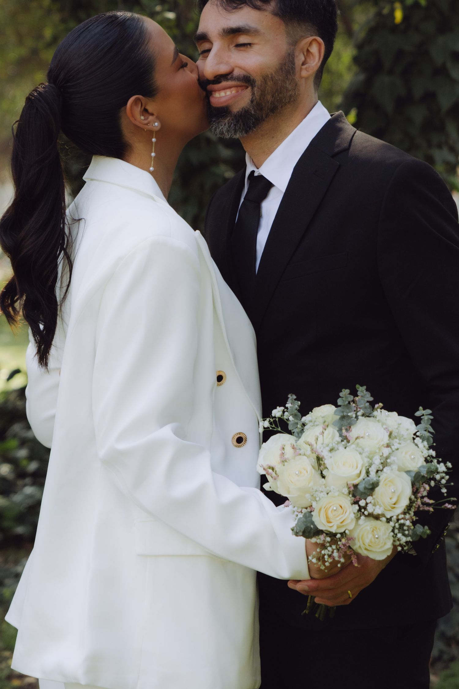 Bride in white blazer kissing grooms cheek while holding a bouquet of white roses outdoors.