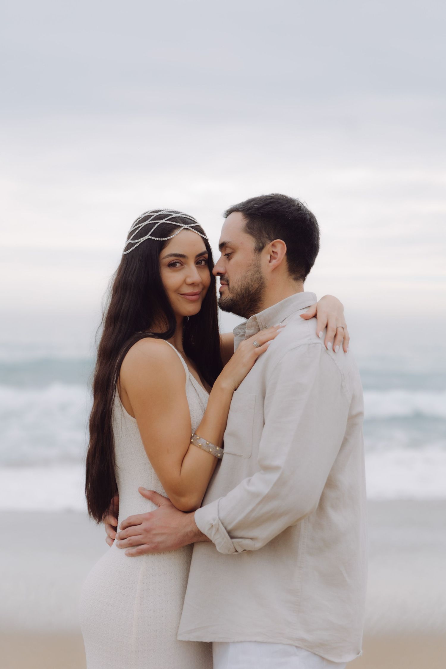 Elegant couple embracing on a cloudy beach, woman in white dress with headpiece looking at camera.
