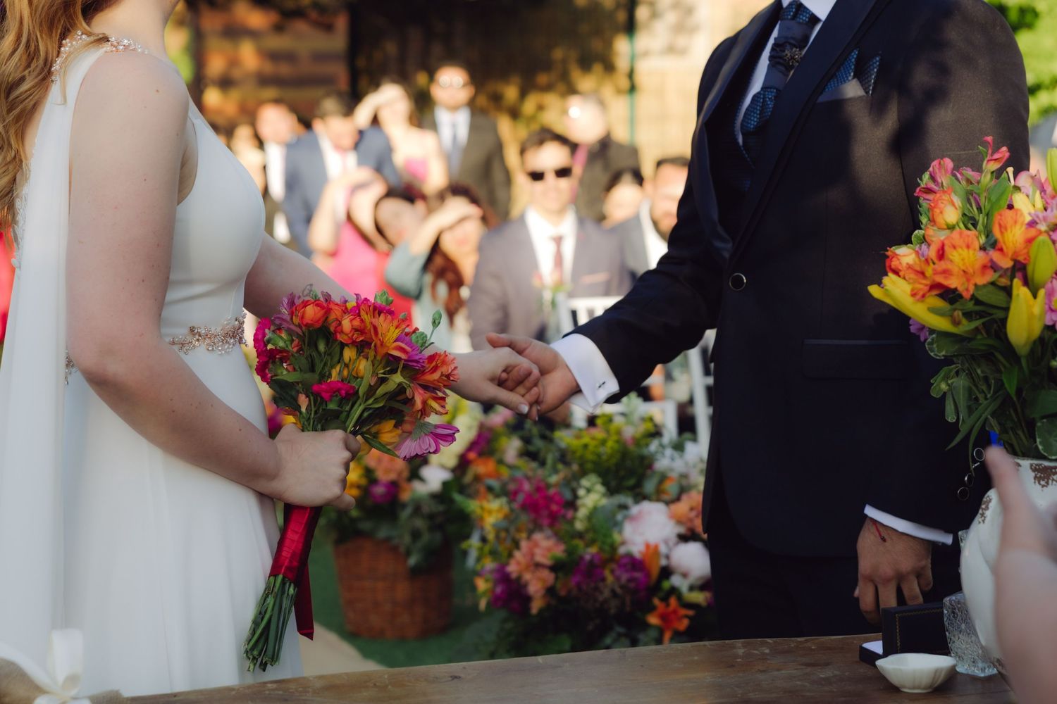 Bride and groom exchange rings at outdoor wedding ceremony surrounded by colorful floral arrangements.