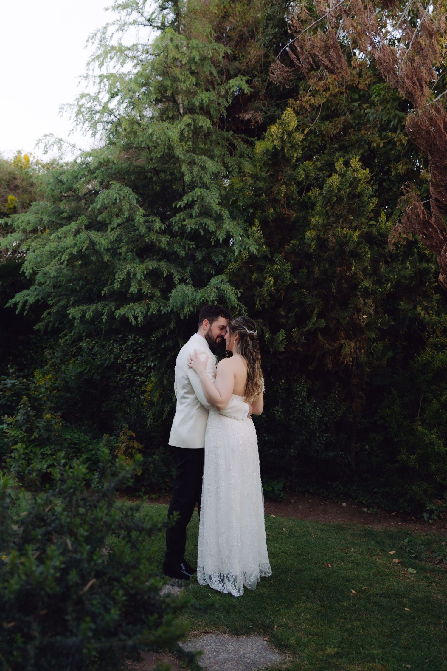 Bride and groom sharing a kiss in a lush green garden surrounded by tall trees at dusk.