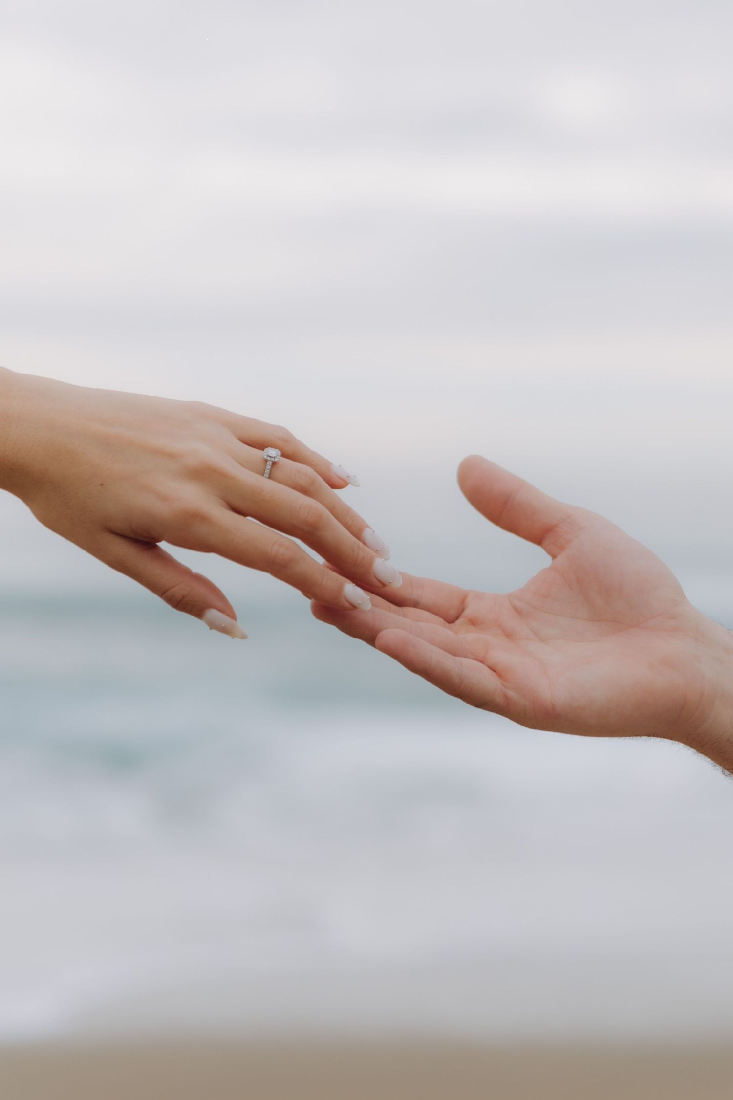 Close-up of two hands reaching toward each other, woman wearing a diamond engagement ring with ocean backdrop.