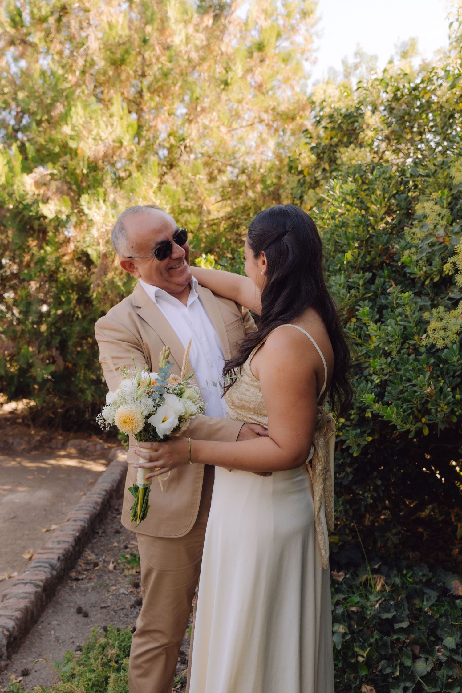 Smiling bald groom in tan suit holds bouquet while bride in ivory gown embraces him outdoors.