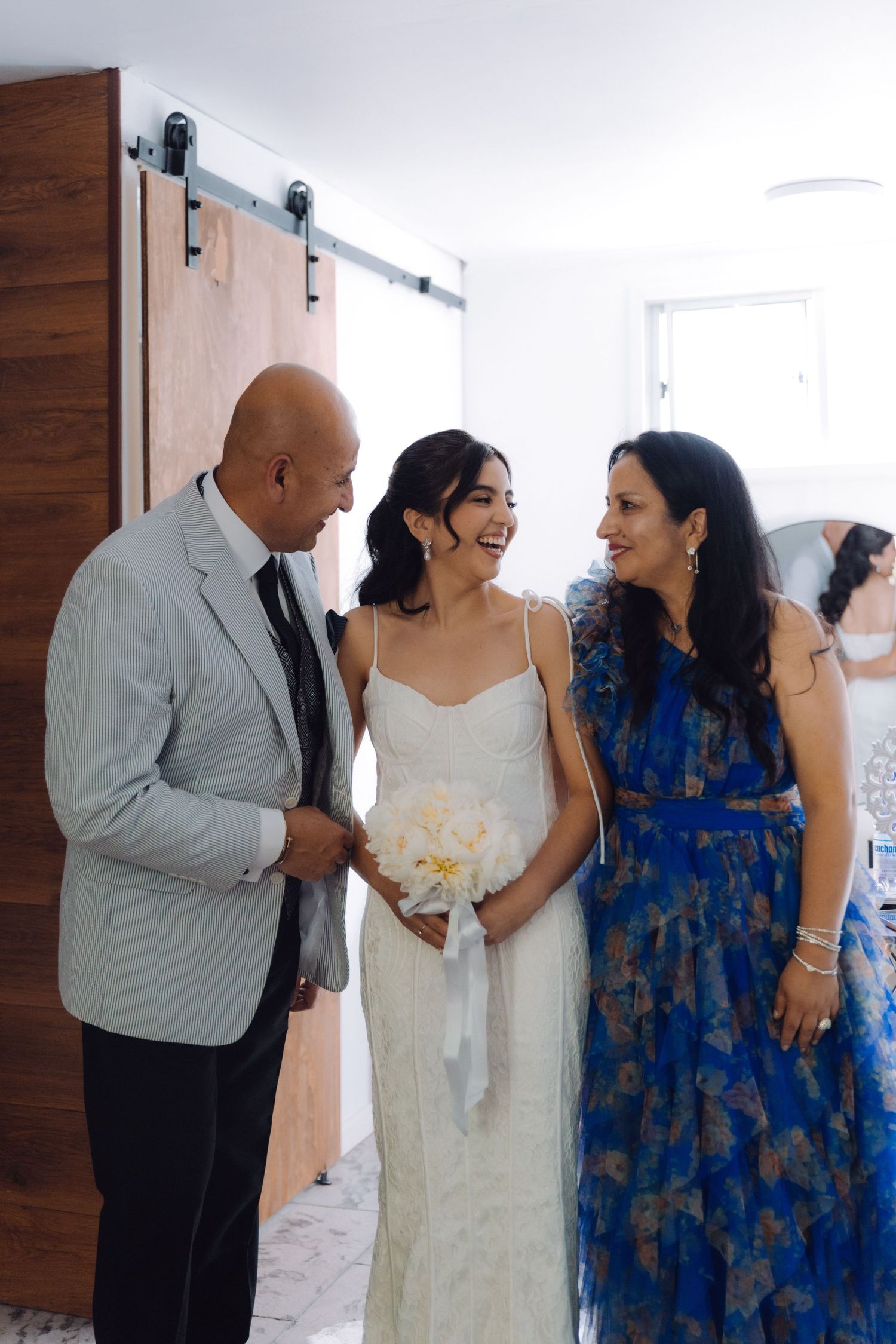 Bride in white lace dress with bouquet sharing a joyful moment with parents before wedding ceremony.