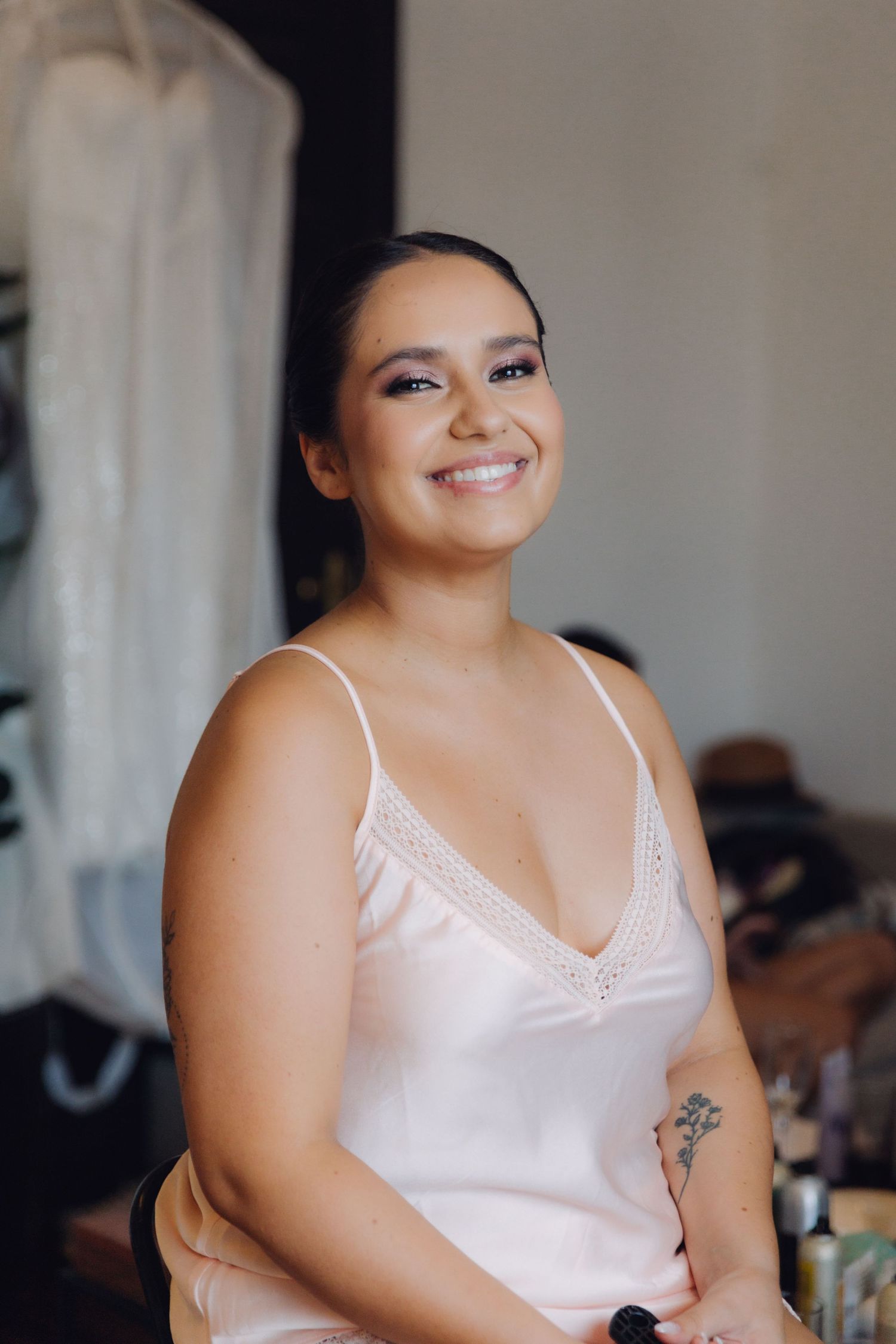 Smiling bride in pink satin slip dress sits during wedding preparations with bridal gown visible in background.