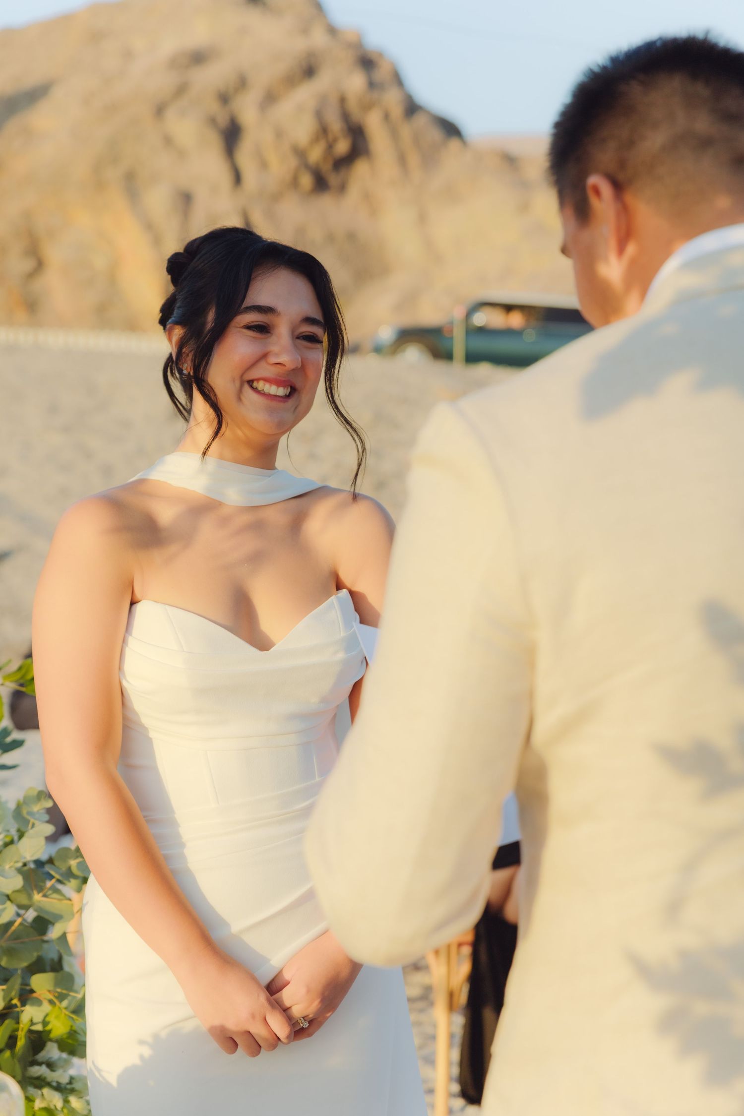 Radiant bride in white strapless gown smiles at groom during outdoor desert wedding ceremony.