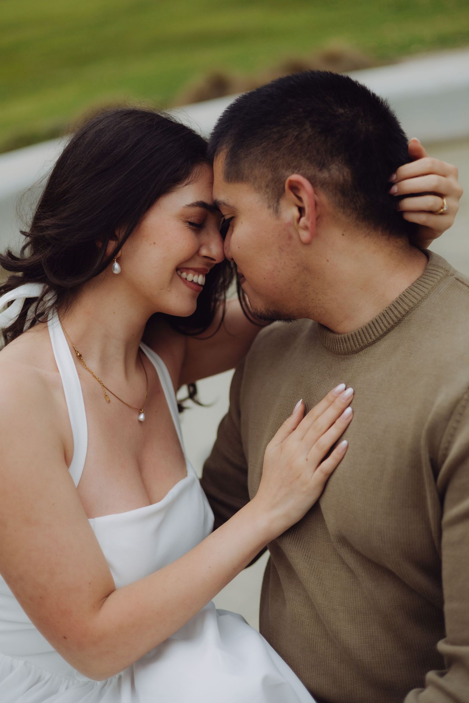 Smiling couple touching foreheads in a tender romantic moment outdoors, woman in white dress holding mans neck.