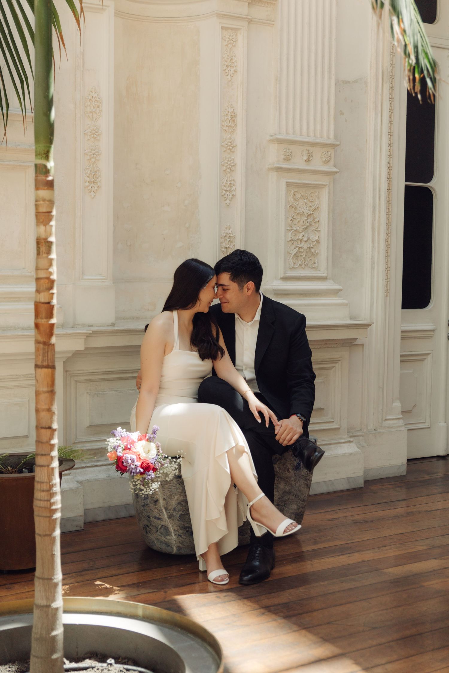 Couple sharing an intimate moment seated together in an elegant white ornate interior with palm trees.