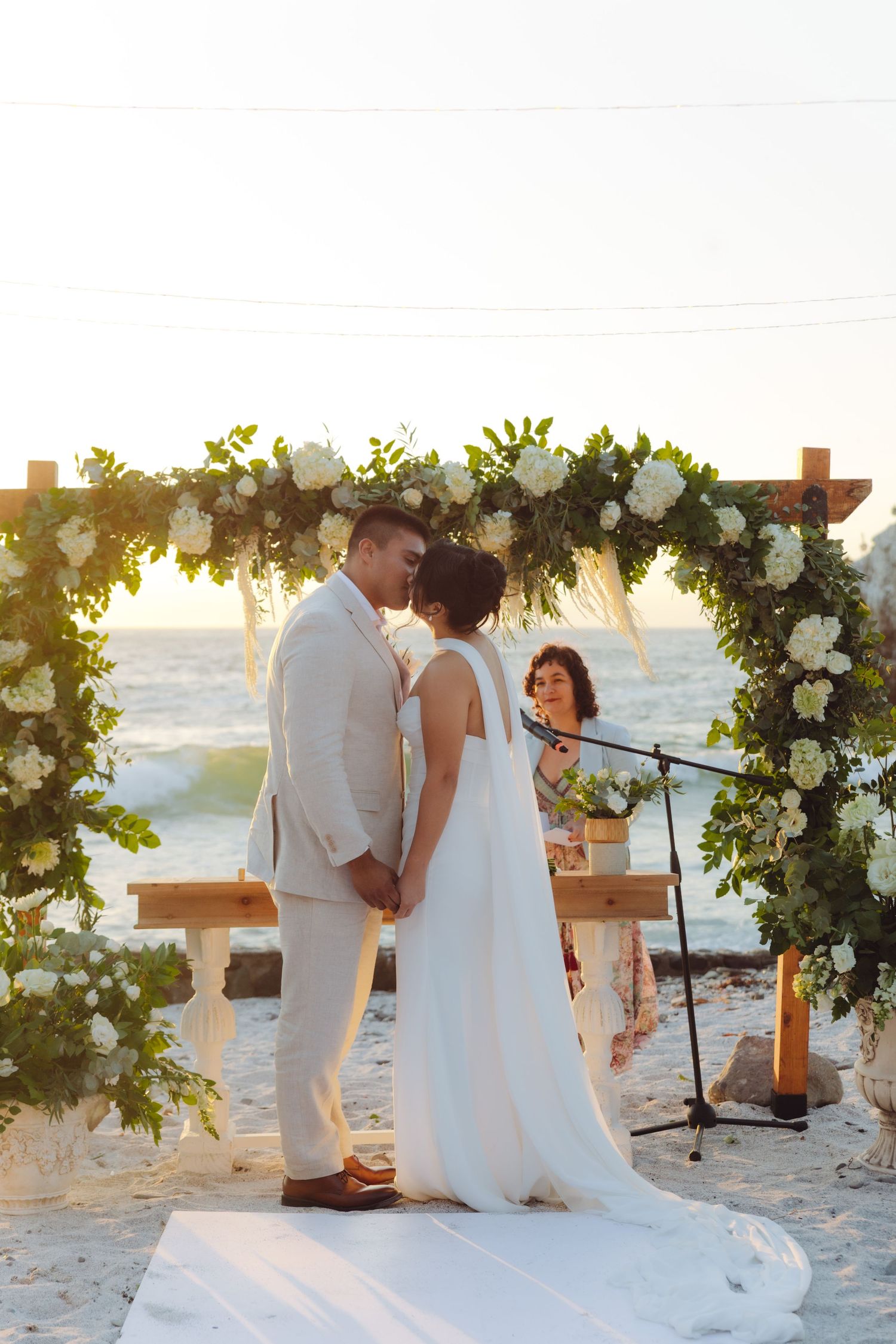 Bride and groom share first kiss under floral arch at sunset beach wedding ceremony.