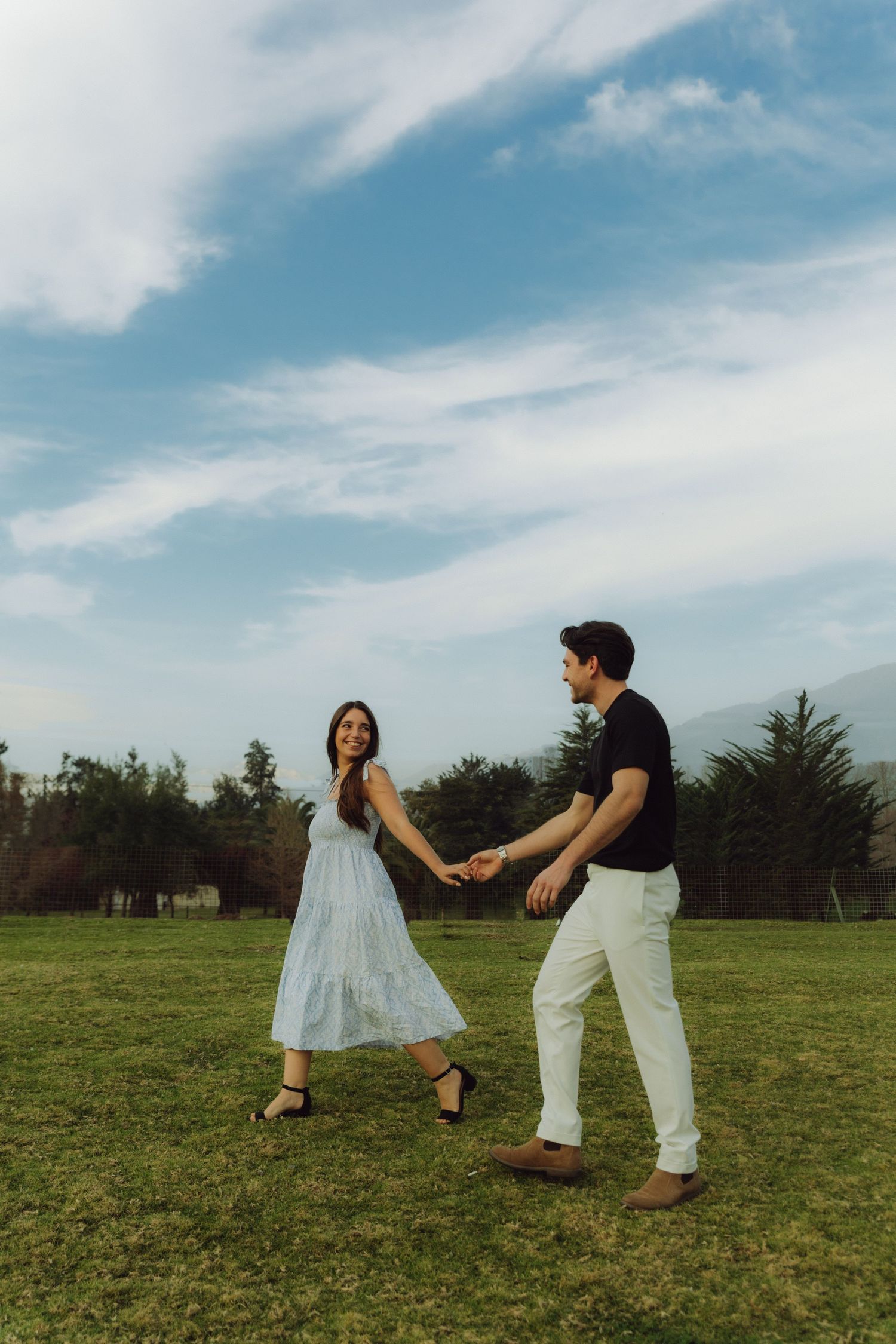 Happy couple holding hands and dancing playfully in an open green field under a blue sky.