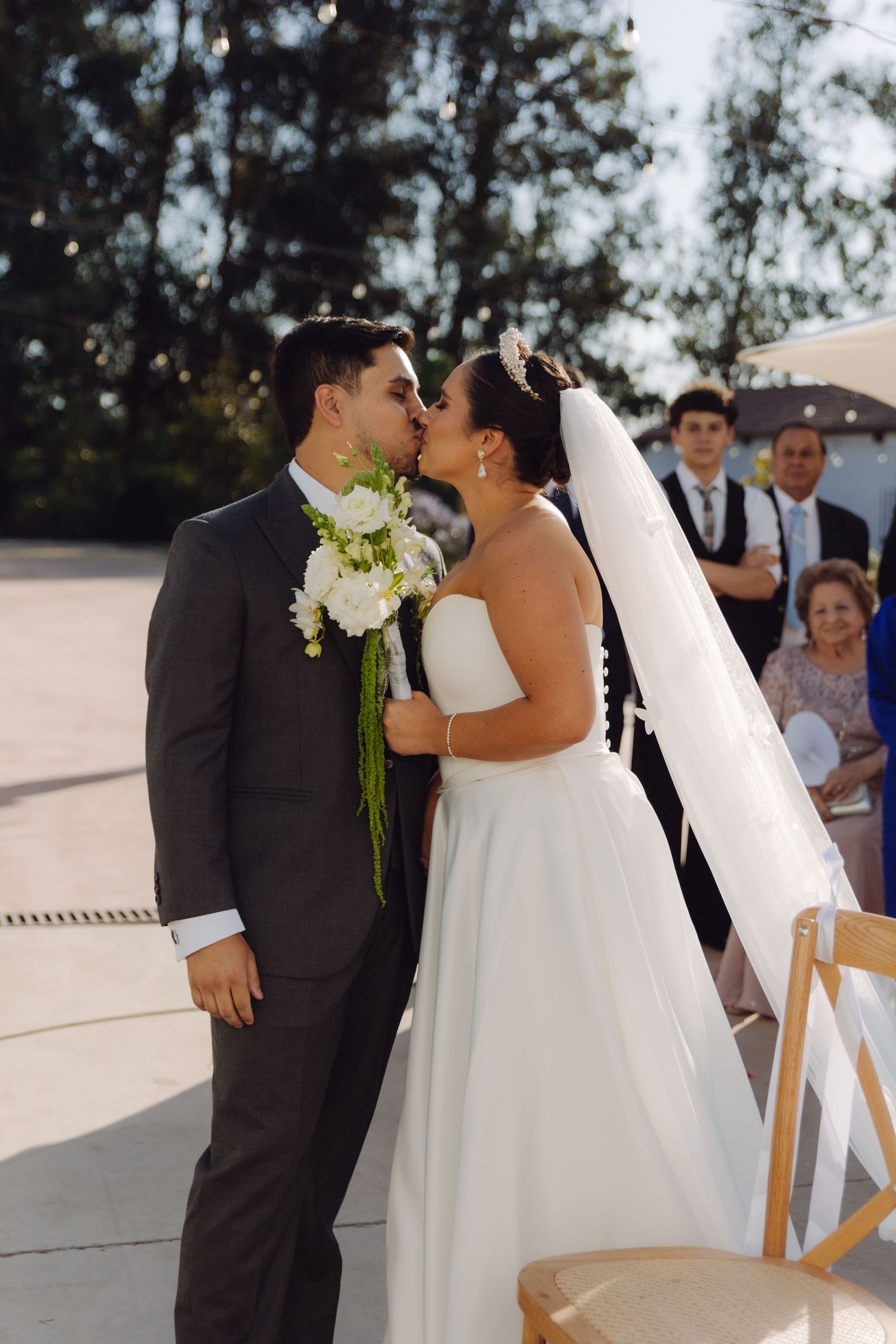 Bride and groom share their first kiss at outdoor wedding ceremony, bride holding white floral bouquet with veil flowing.