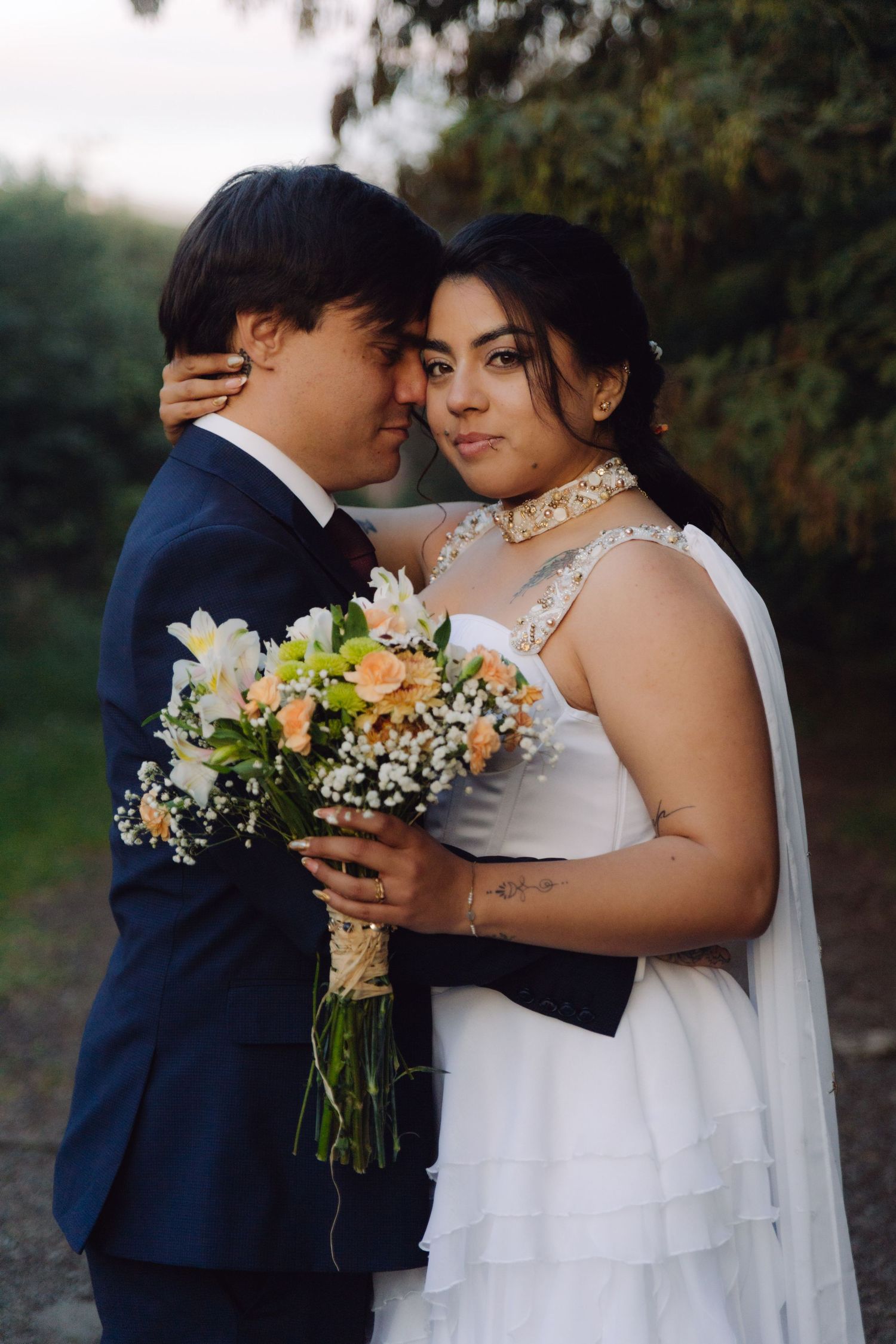 Bride holding colorful bouquet embracing groom in navy suit while looking at camera in outdoor setting.