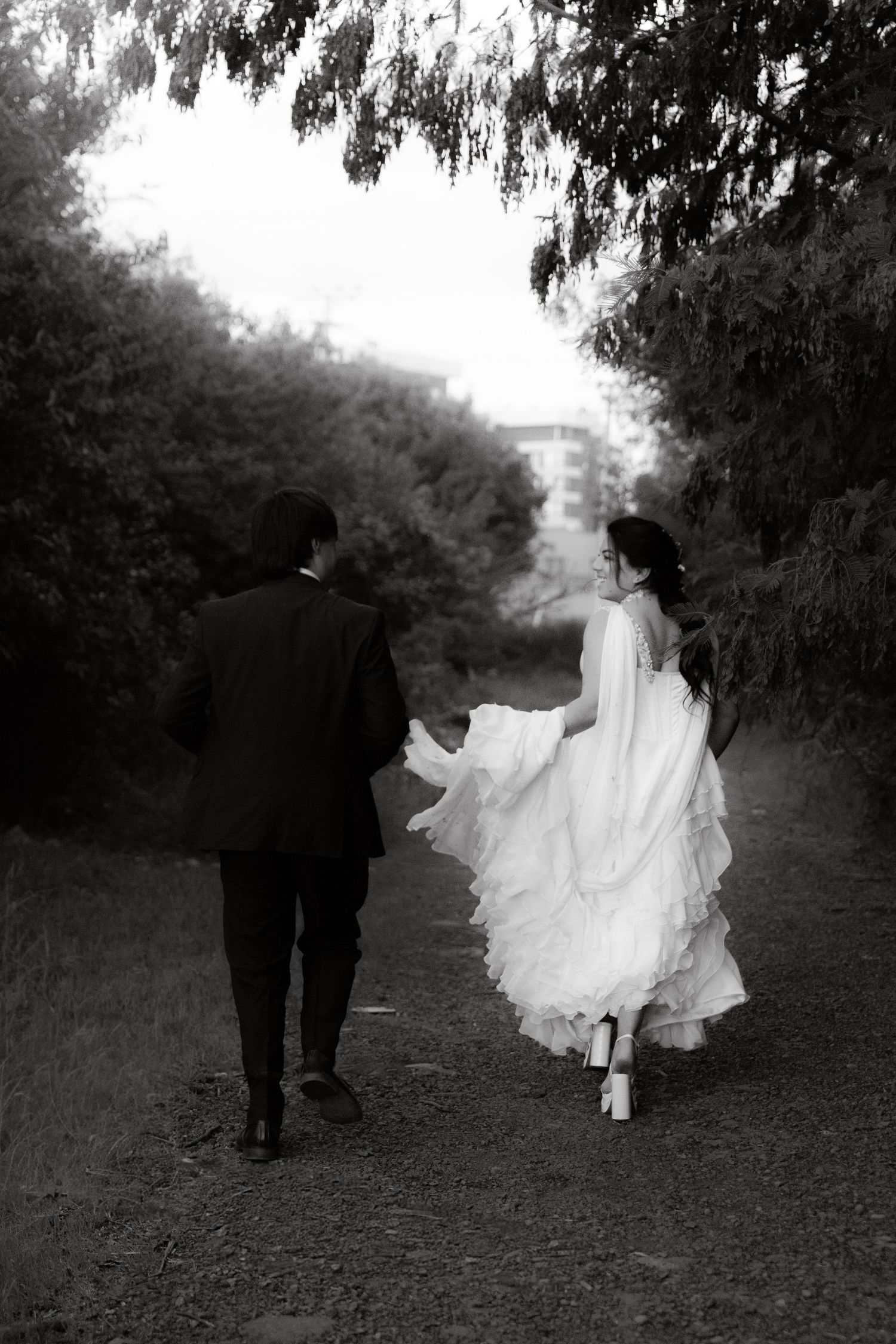 Black and white photo of bride and groom walking hand in hand down a tree-lined path.
