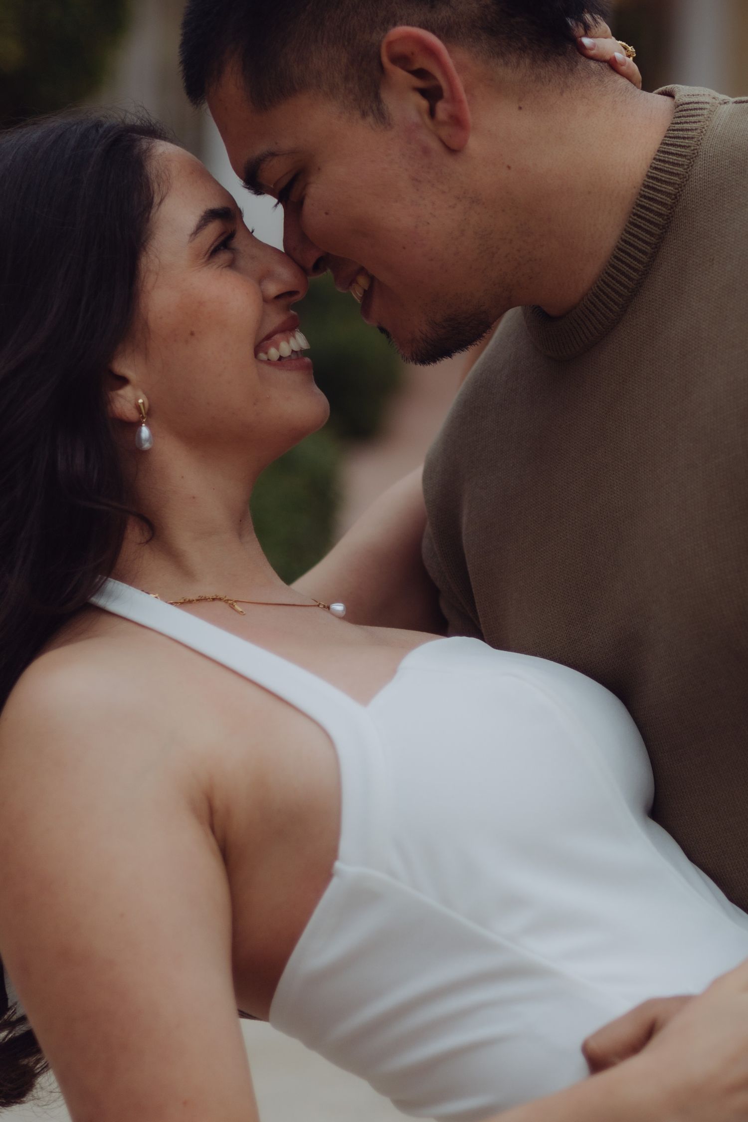 Smiling pregnant woman in white dress shares a romantic moment with her partner outdoors.