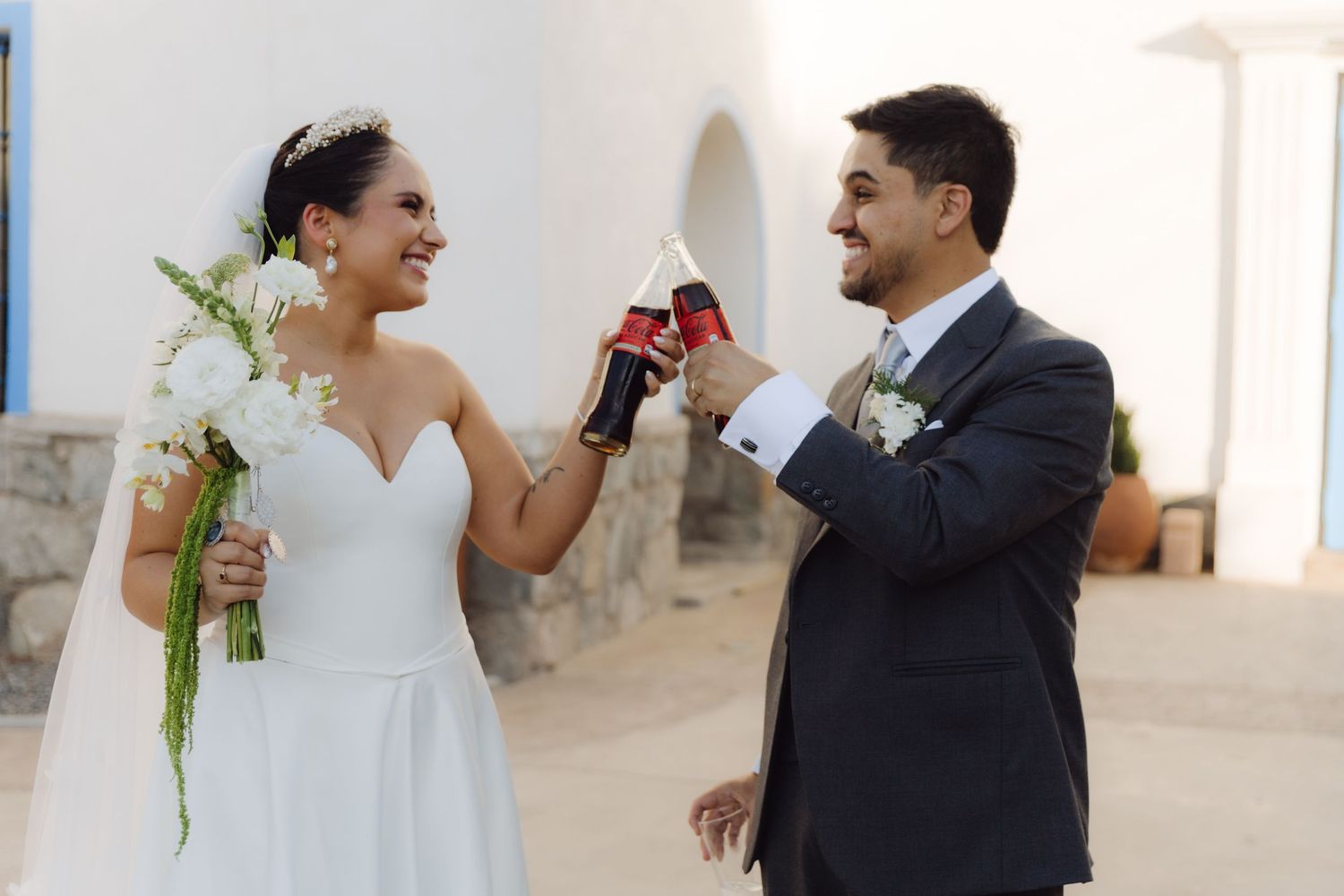 Joyful bride and groom toast with Coca-Cola bottles outside a white building, bride holding white wedding bouquet.