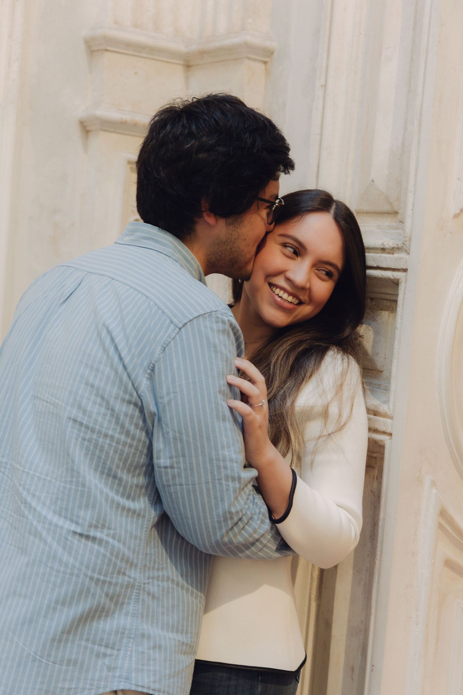 Man kissing smiling woman on cheek against ornate stone wall outdoors.
