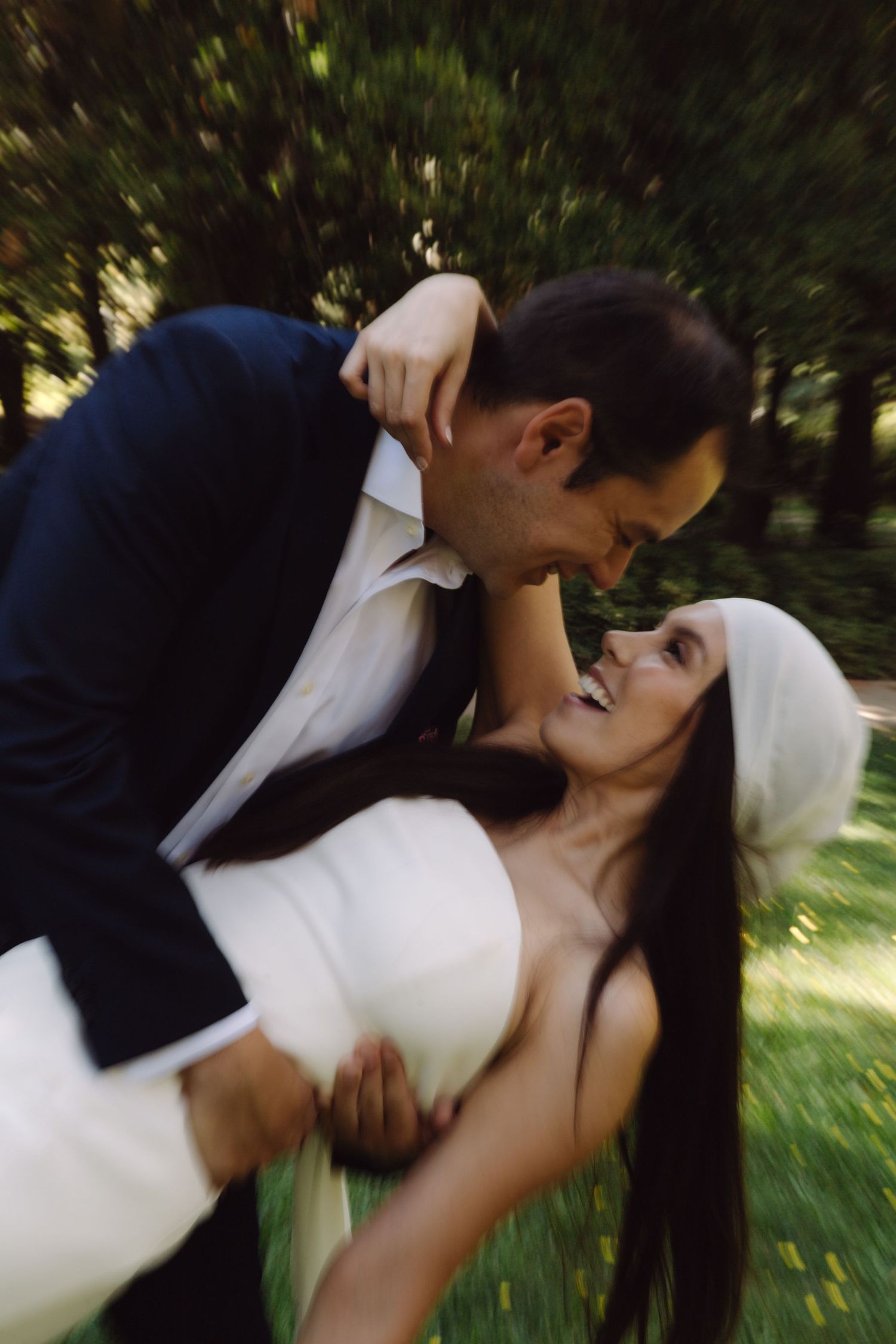 Groom dipping smiling bride in white dress and veil outdoors in lush green park setting.