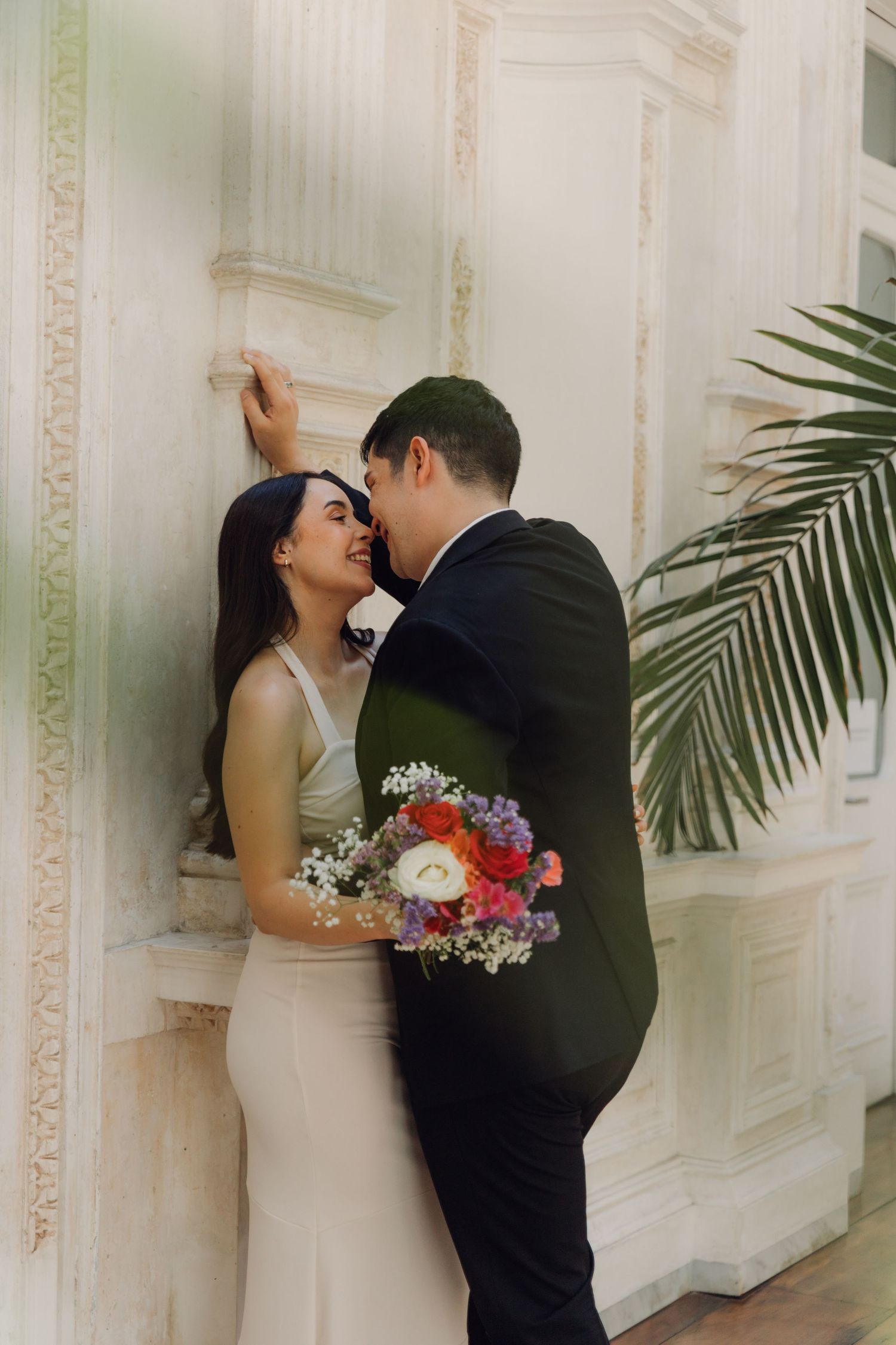 Bride and groom sharing a tender moment against ornate white wall, bride holding colorful bouquet.
