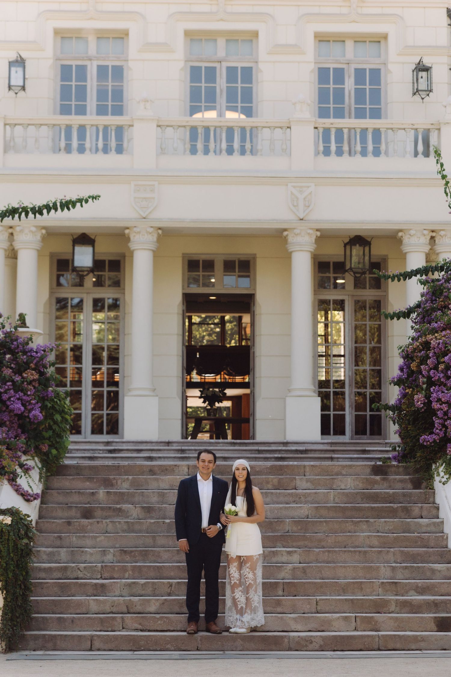 Newlywed couple stands on grand steps of an elegant building adorned with purple flowers.