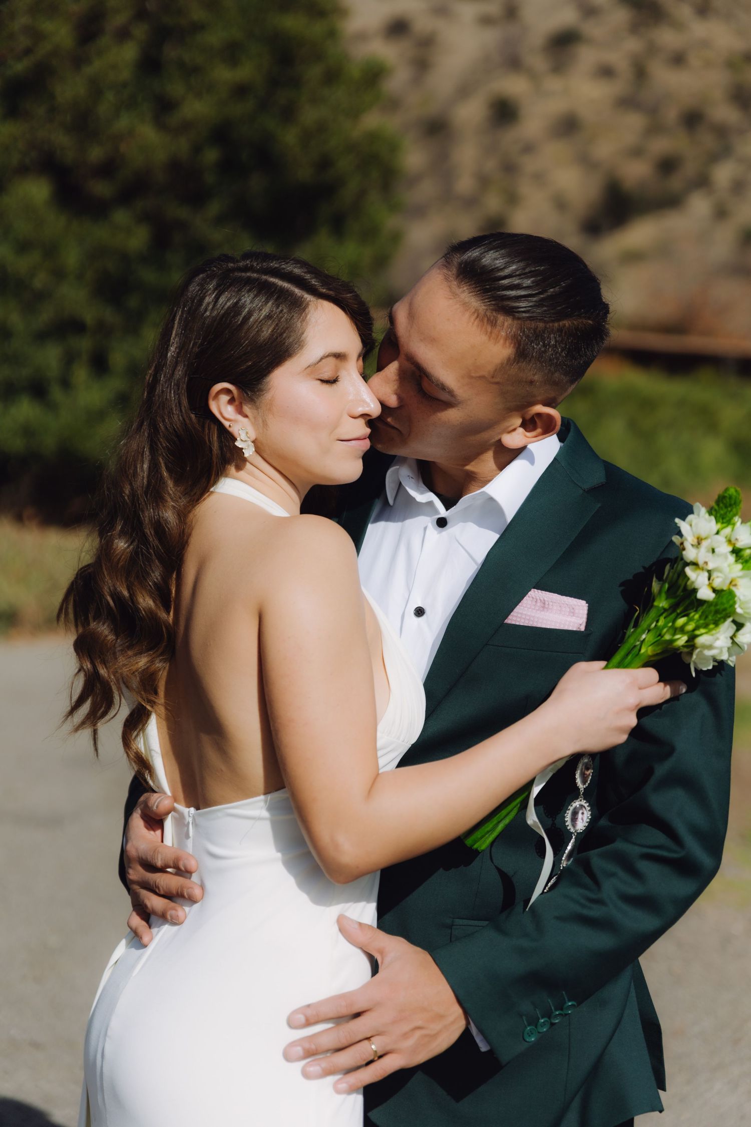Groom in green suit kissing bride on cheek while embracing outdoors, bride holding white flower bouquet.