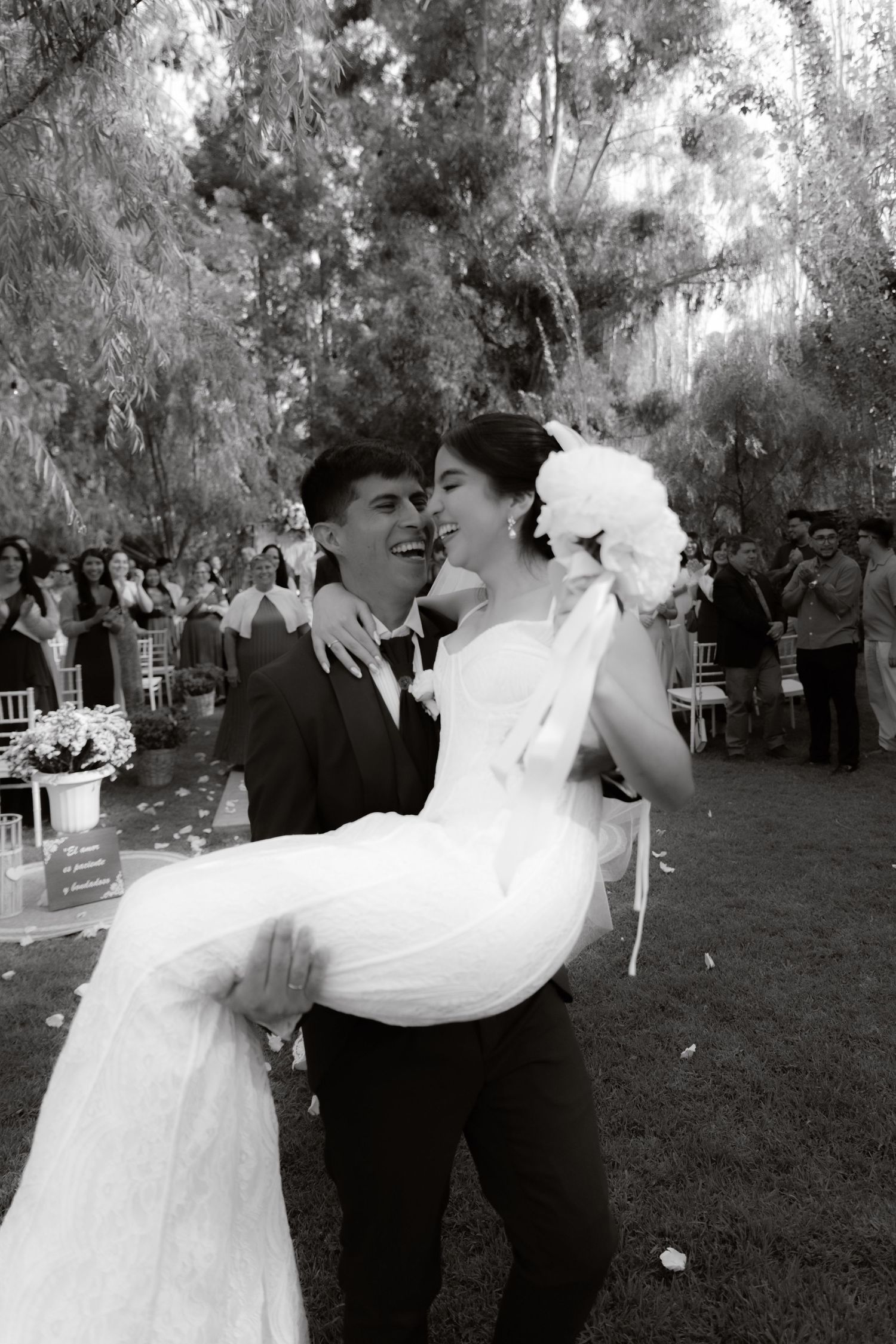 Joyful groom carries laughing bride in white wedding gown outdoors in black and white wedding photo.