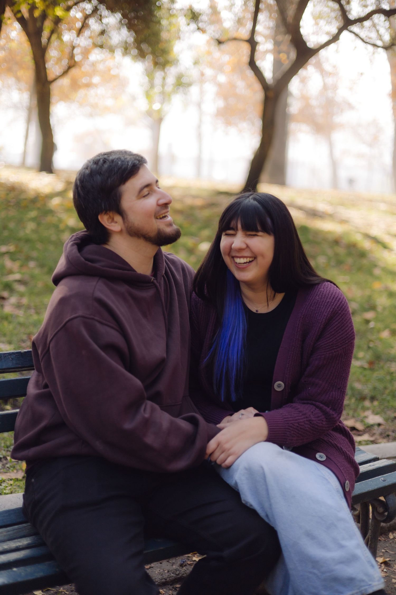Happy couple laughing together on a park bench in autumn, woman with blue highlighted hair in purple cardigan.