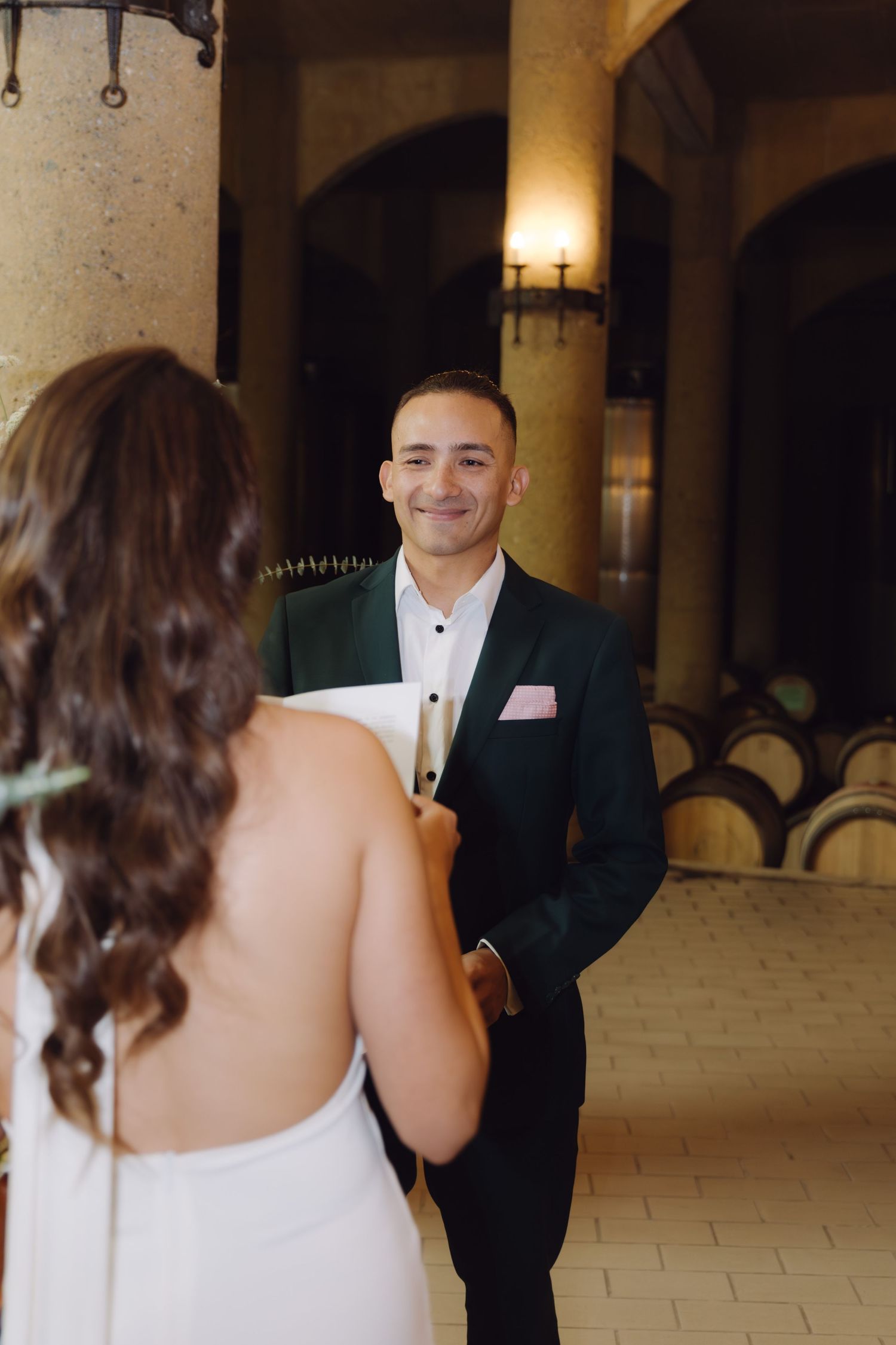 Smiling groom in green tuxedo holds brides hands during intimate first look ceremony in rustic wine cellar.