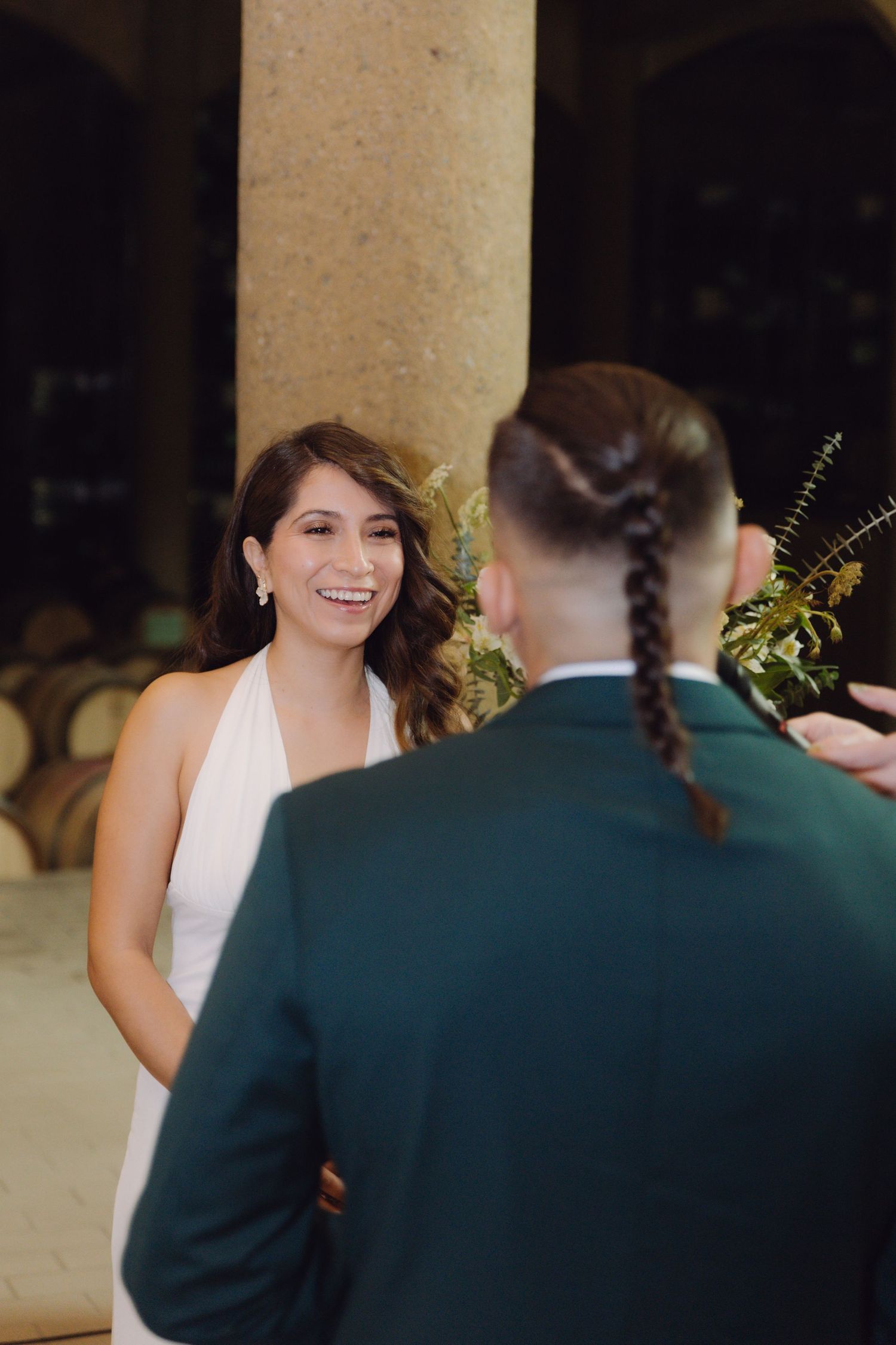 Joyful bride in white halter dress smiling at groom during wedding ceremony in elegant barrel room venue.