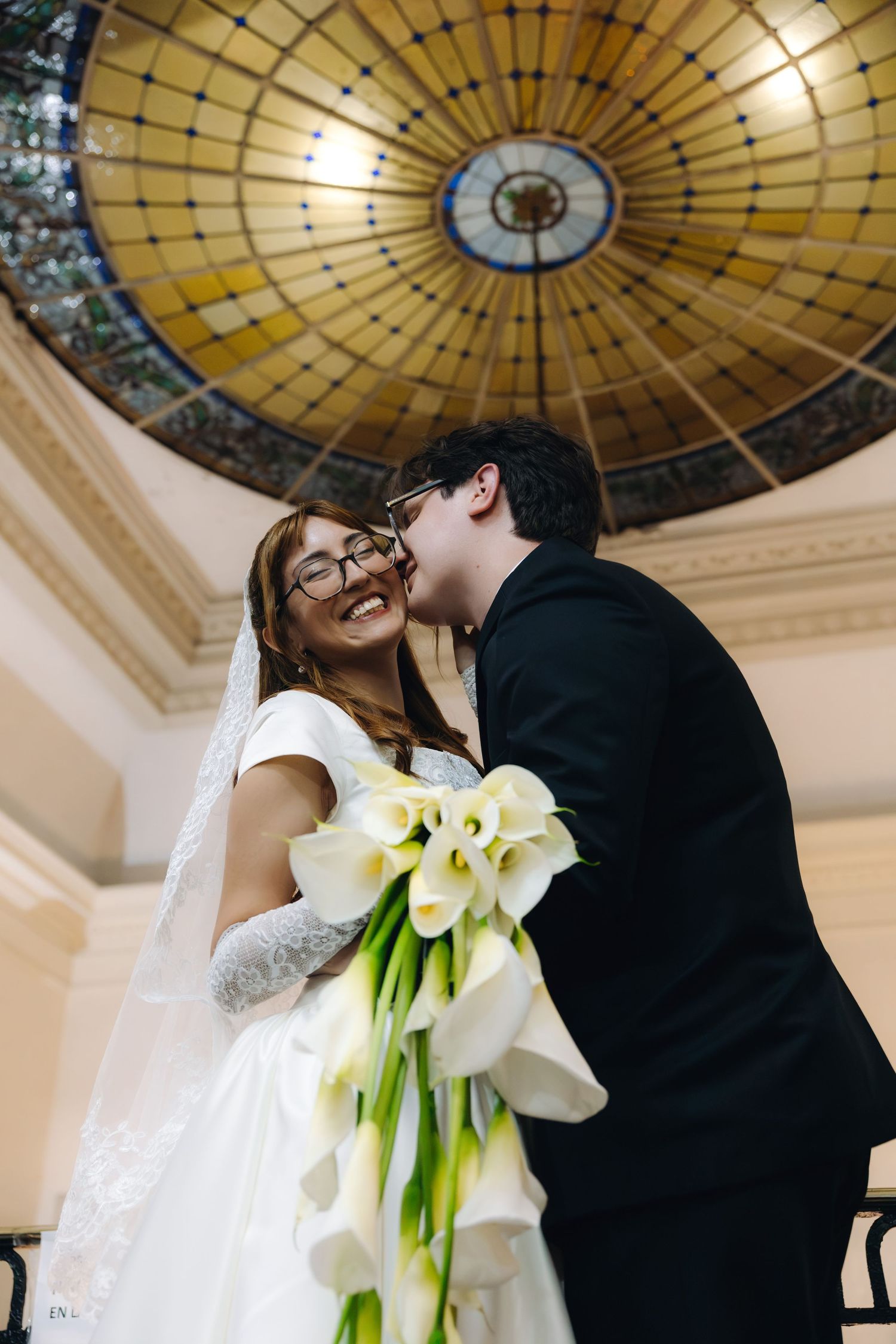 Groom kisses smiling bride wearing glasses beneath ornate stained glass dome holding calla lilies.