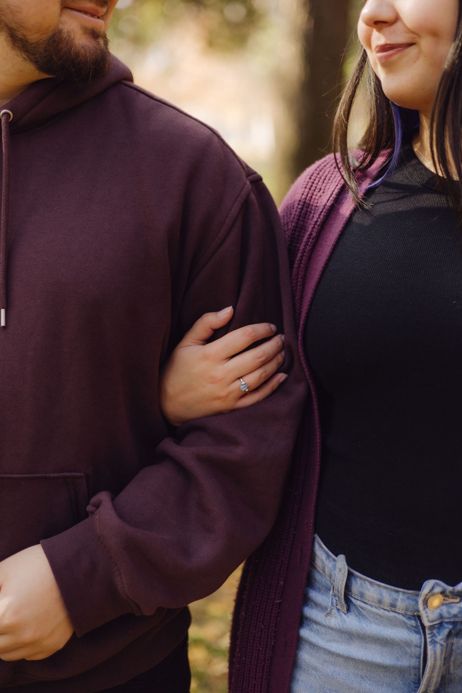 Close-up of couple embracing outdoors, woman showing engagement ring while holding mans arm in autumn.