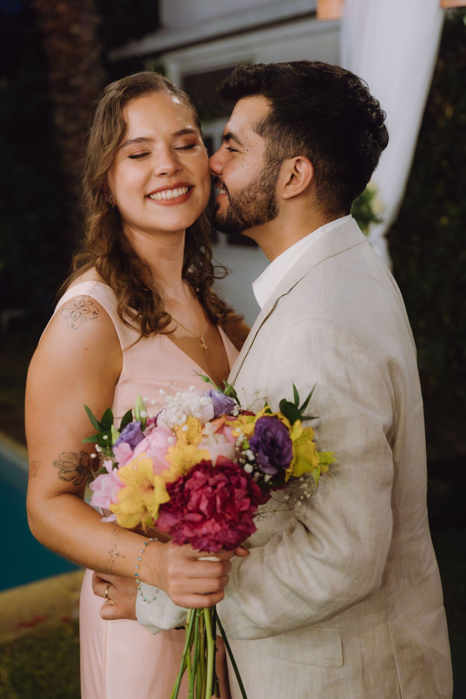 Smiling bride in pink dress holding colorful bouquet as groom in beige suit kisses her cheek.