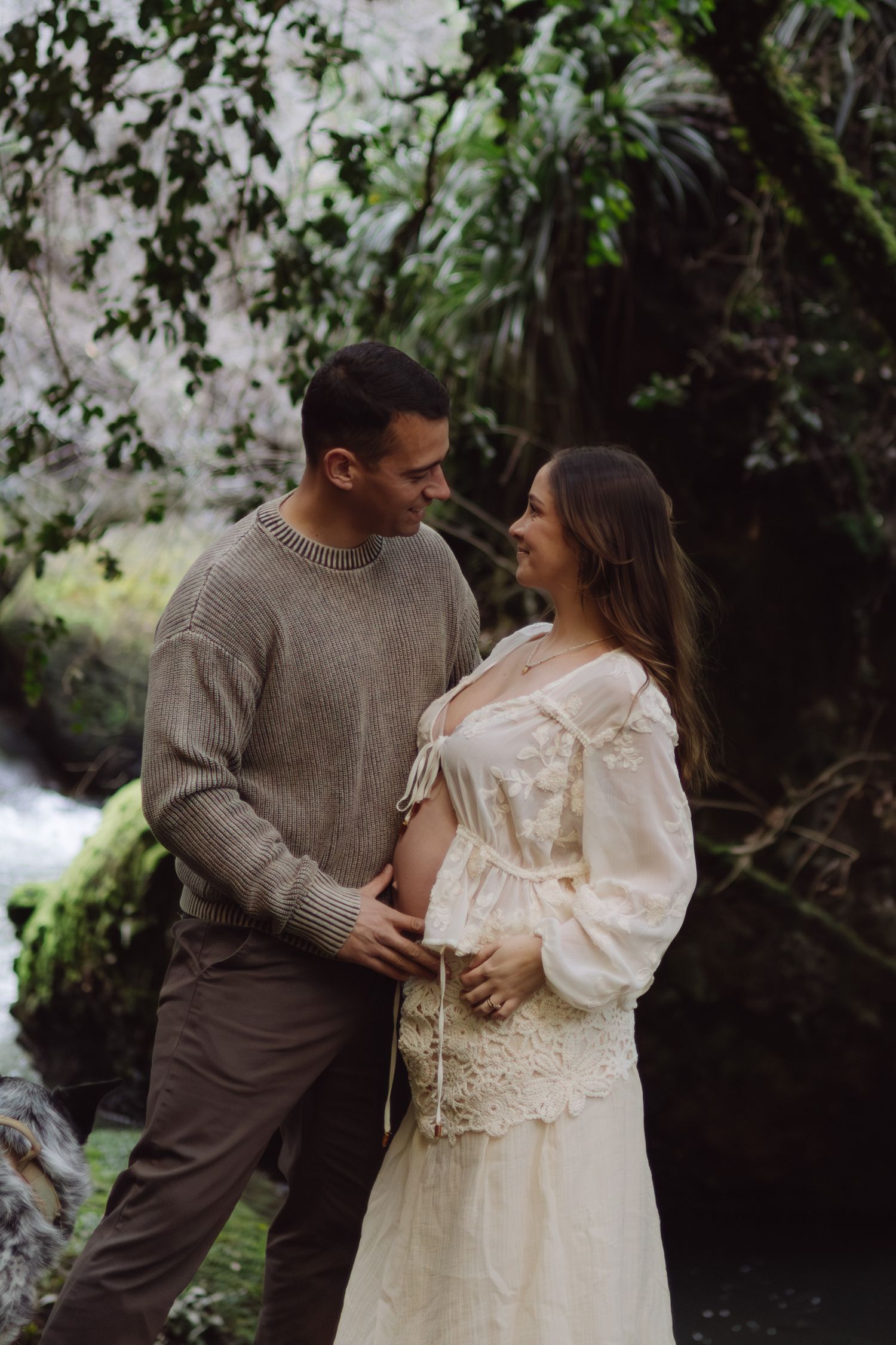 Happy couple touching pregnant belly together near a lush forest waterfall backdrop.