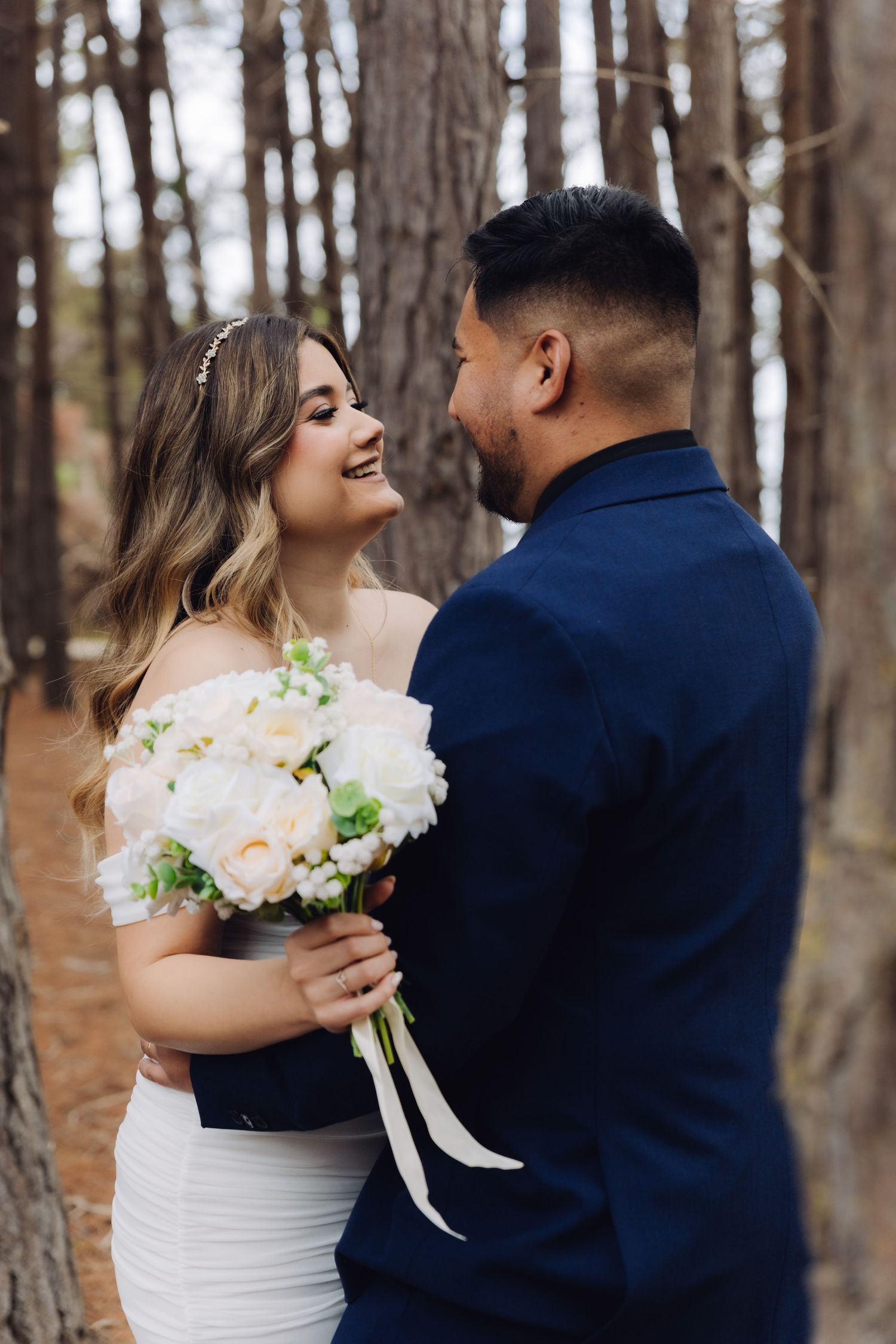 Smiling bride in white dress holding bouquet faces groom in navy suit in pine forest.