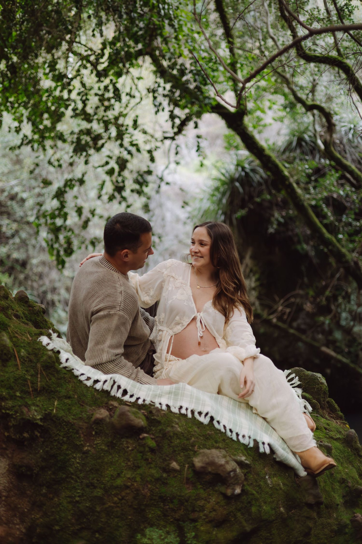 Smiling pregnant couple sitting on a mossy rock in a lush green forest setting.