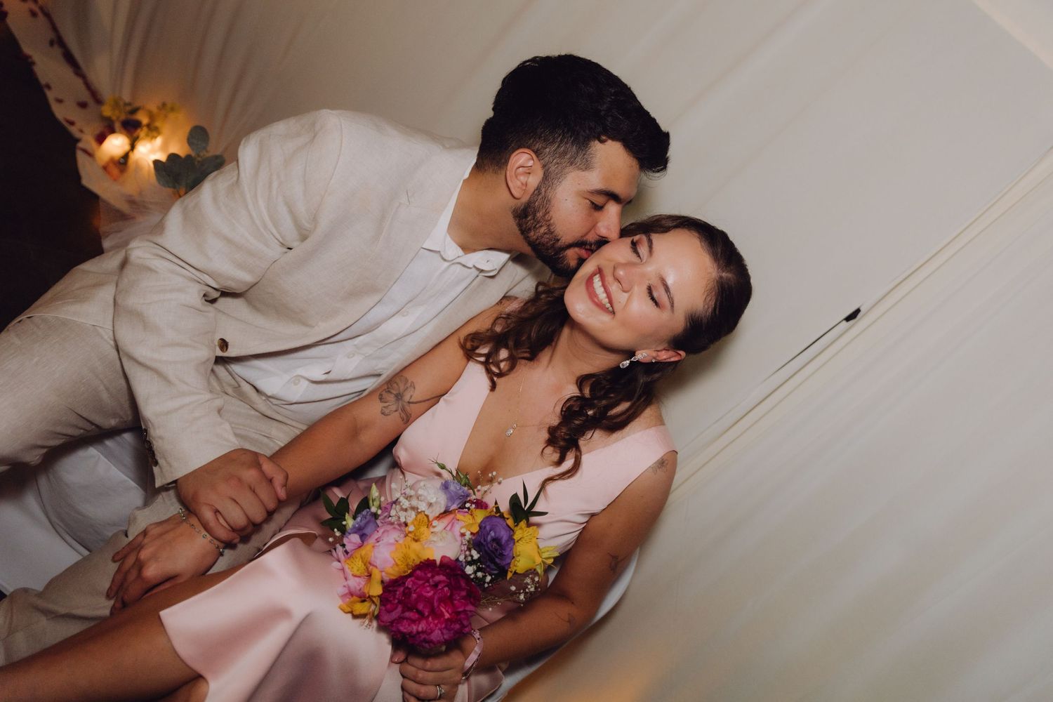 Couple sharing a joyful intimate moment, groom kissing bride holding vibrant floral bouquet.