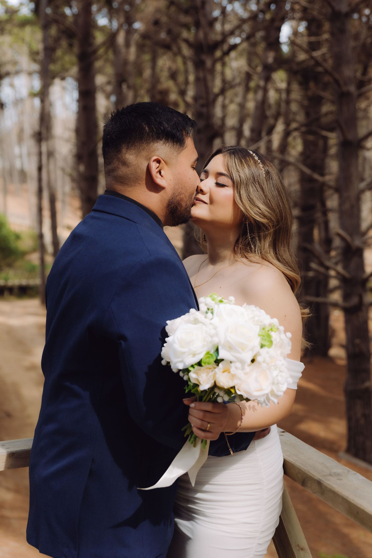 Bride and groom share a kiss in a pine forest, bride holding a white bouquet.