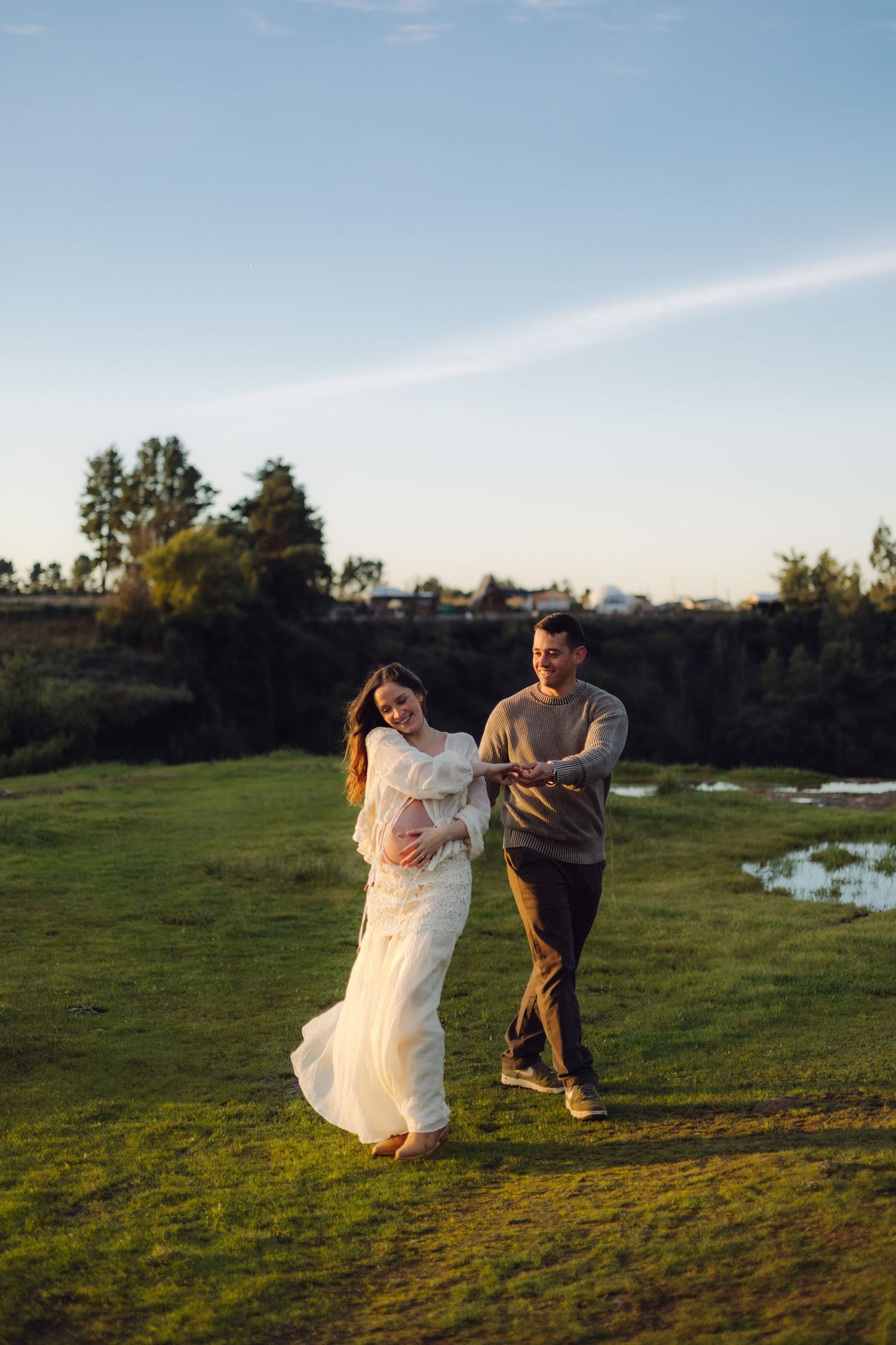 Pregnant woman in flowing white dress walking with partner across a sunny green field at golden hour.