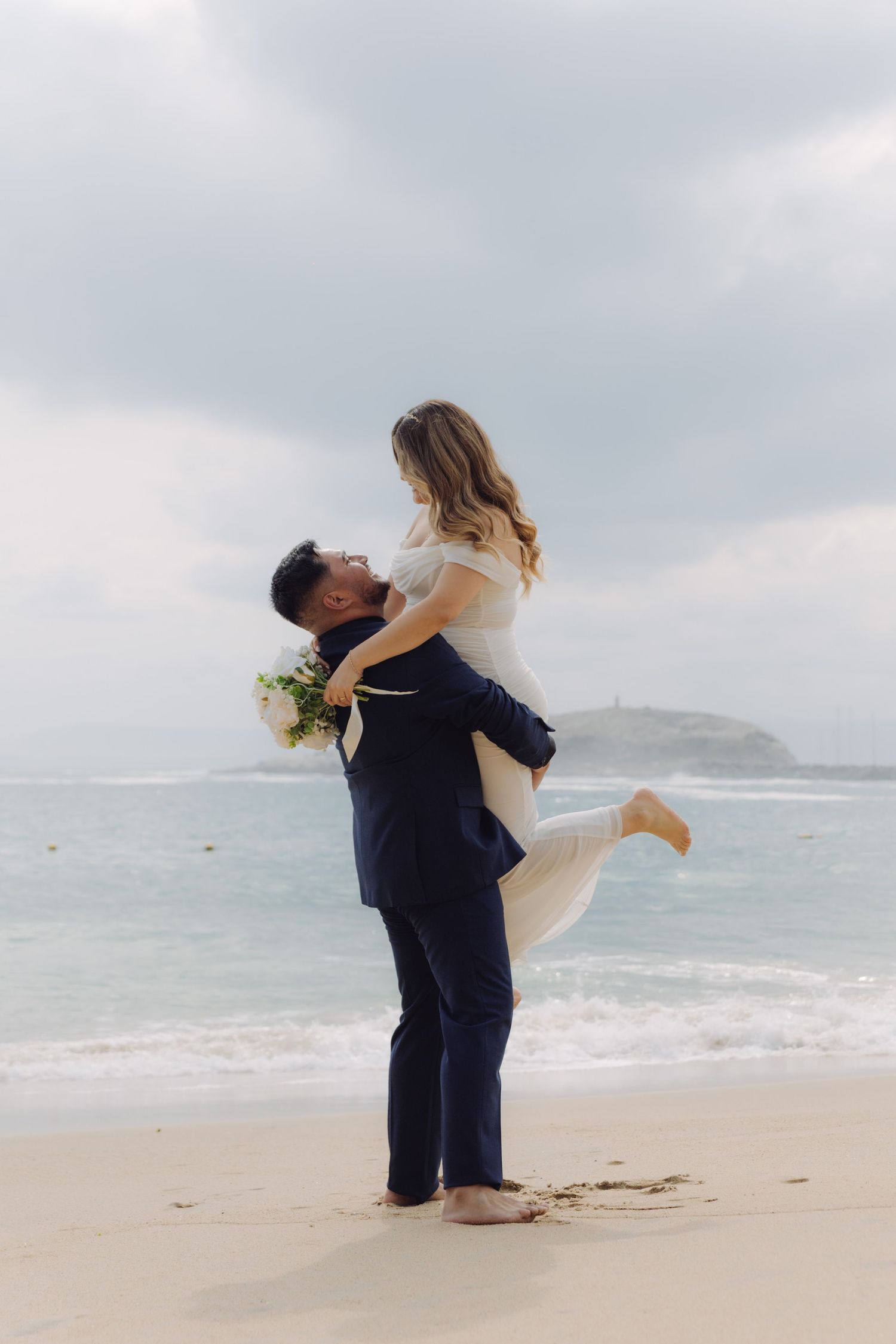 Groom in navy suit lifts bride on sandy beach with ocean waves in background.