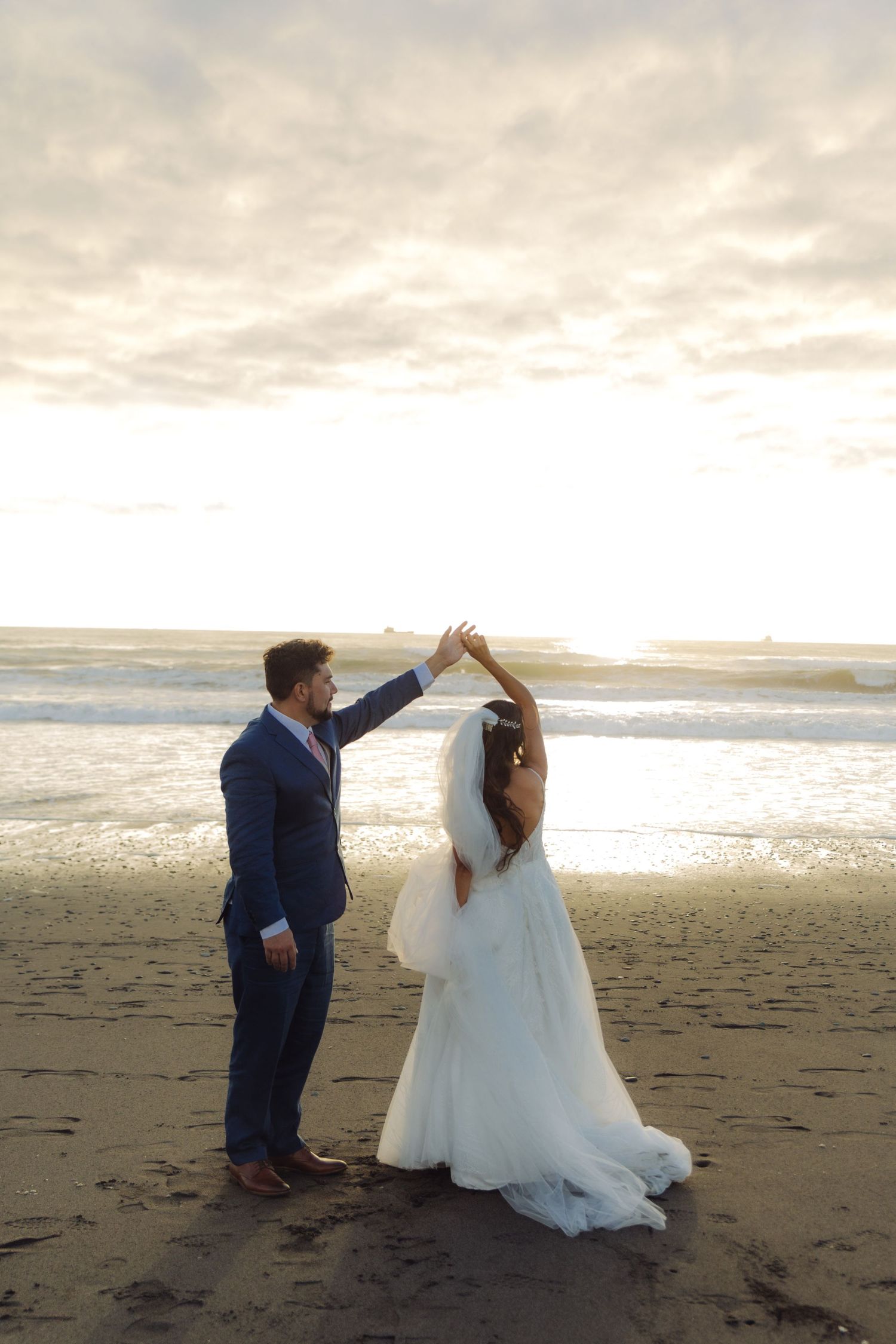 Bride and groom dancing romantically on beach at sunset, groom twirling bride in white gown.