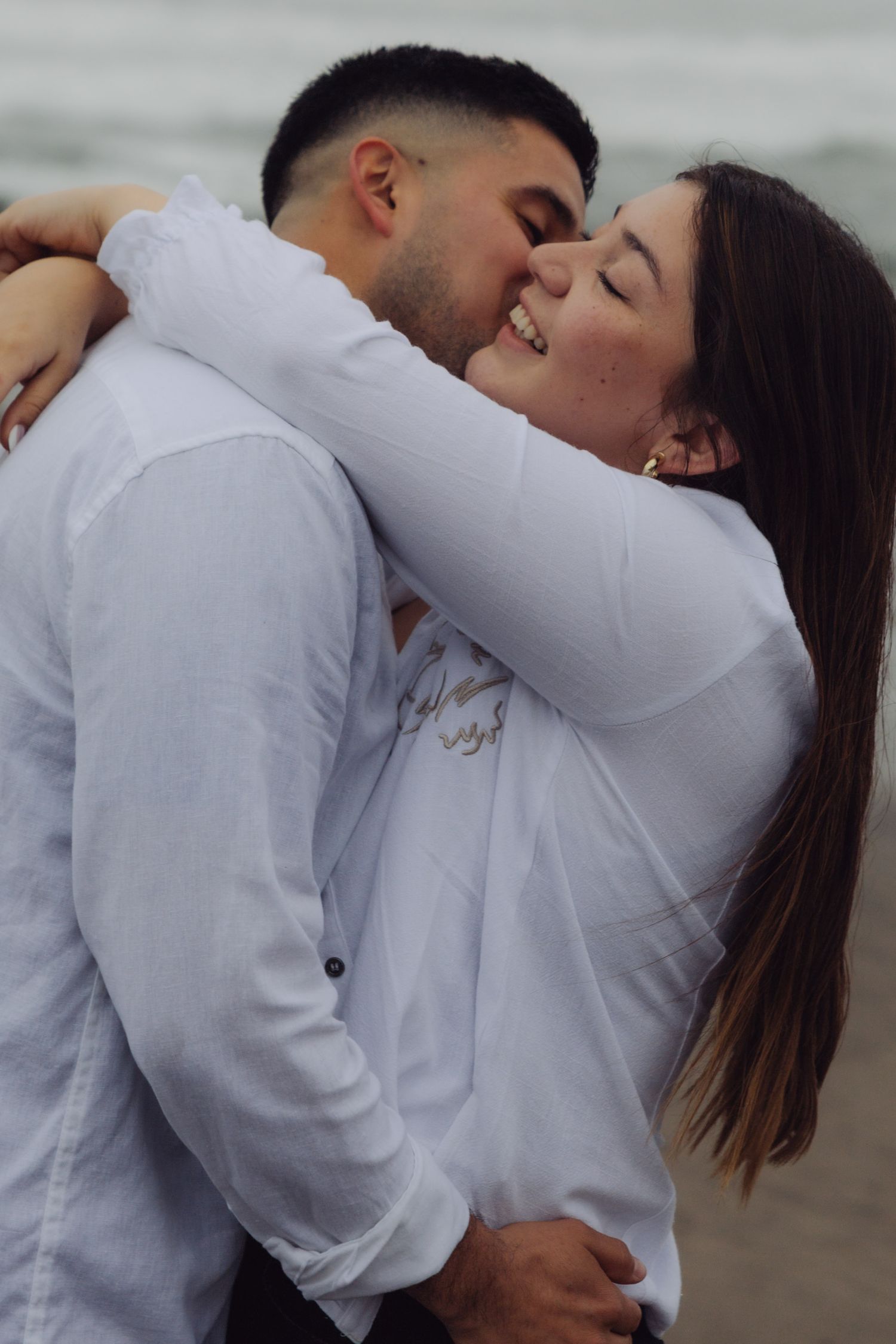 Joyful couple embracing and kissing on a beach, both wearing white outfits with ocean waves behind them.