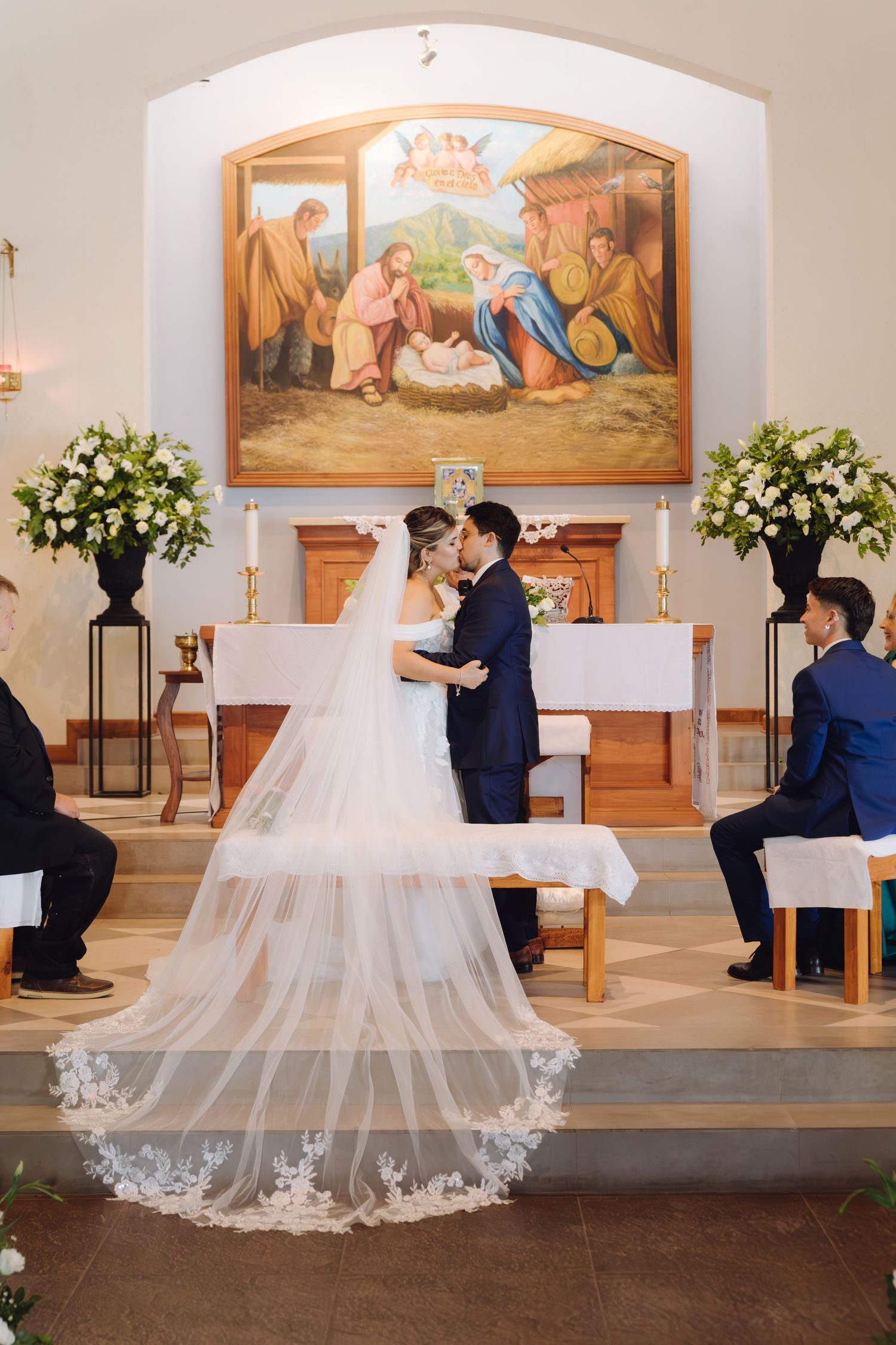 Bride in lace gown with long veil stands with groom at church altar beneath nativity painting.