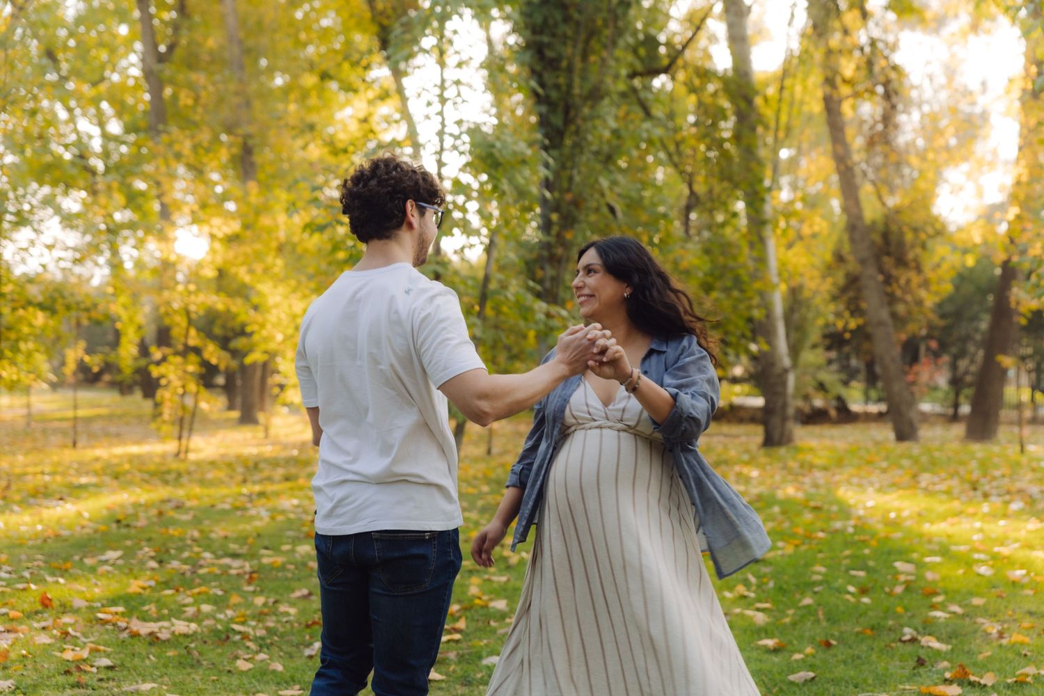 Pregnant woman in a floral dress dances with her partner in a colorful autumn park.