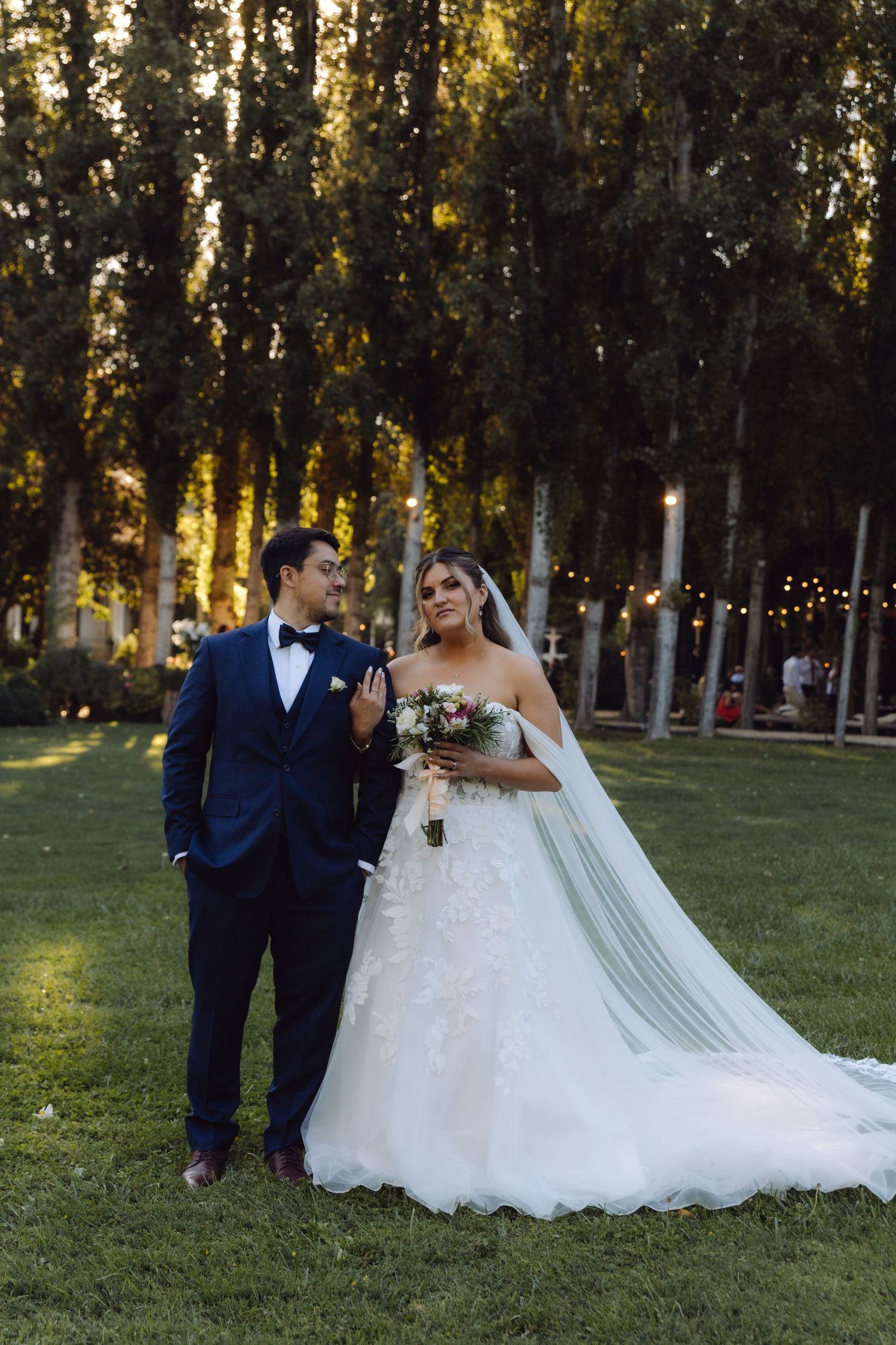 Bride and groom share romantic moment in lush garden at golden hour with string lights.