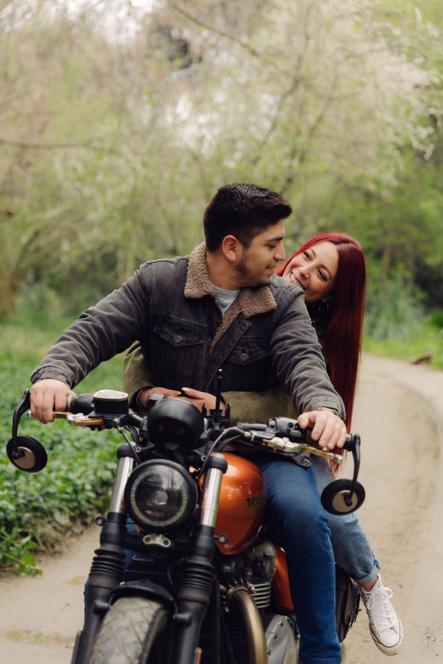 Couple riding a vintage orange motorcycle together through a lush green tree-lined road.
