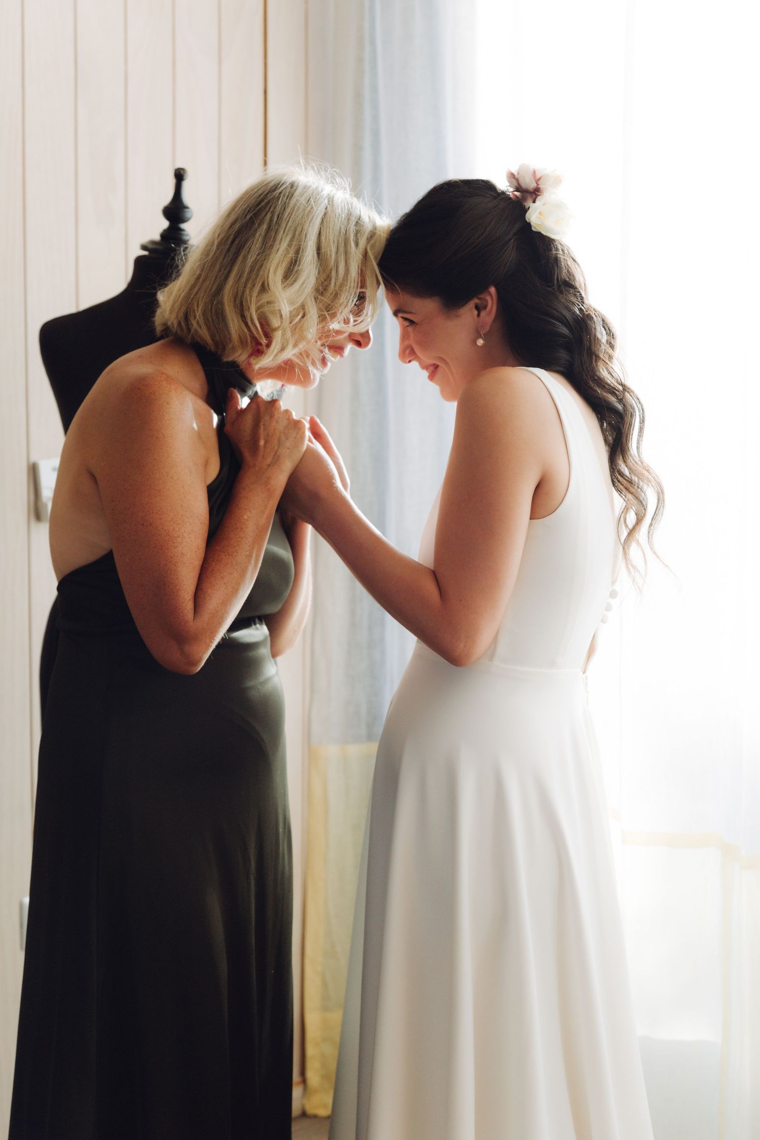 Bride in white dress shares tender moment with older woman in green dress before wedding.