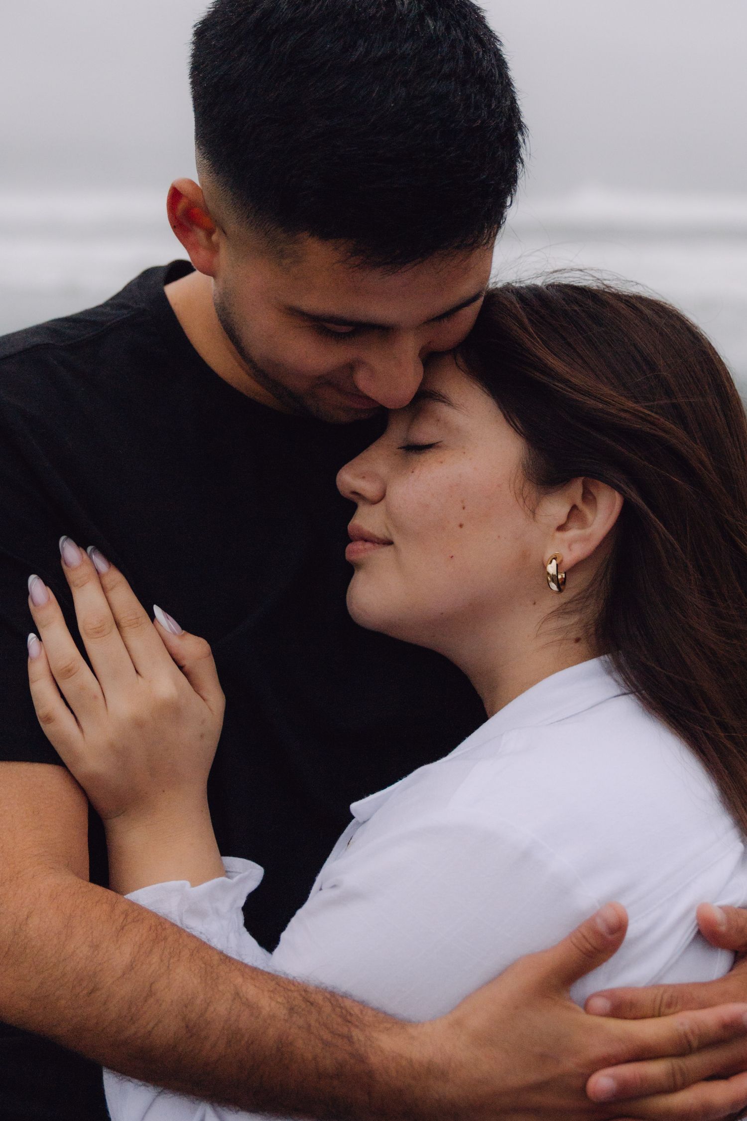 Tender couple moment with man kissing womans forehead while embracing outdoors against a soft grey background.